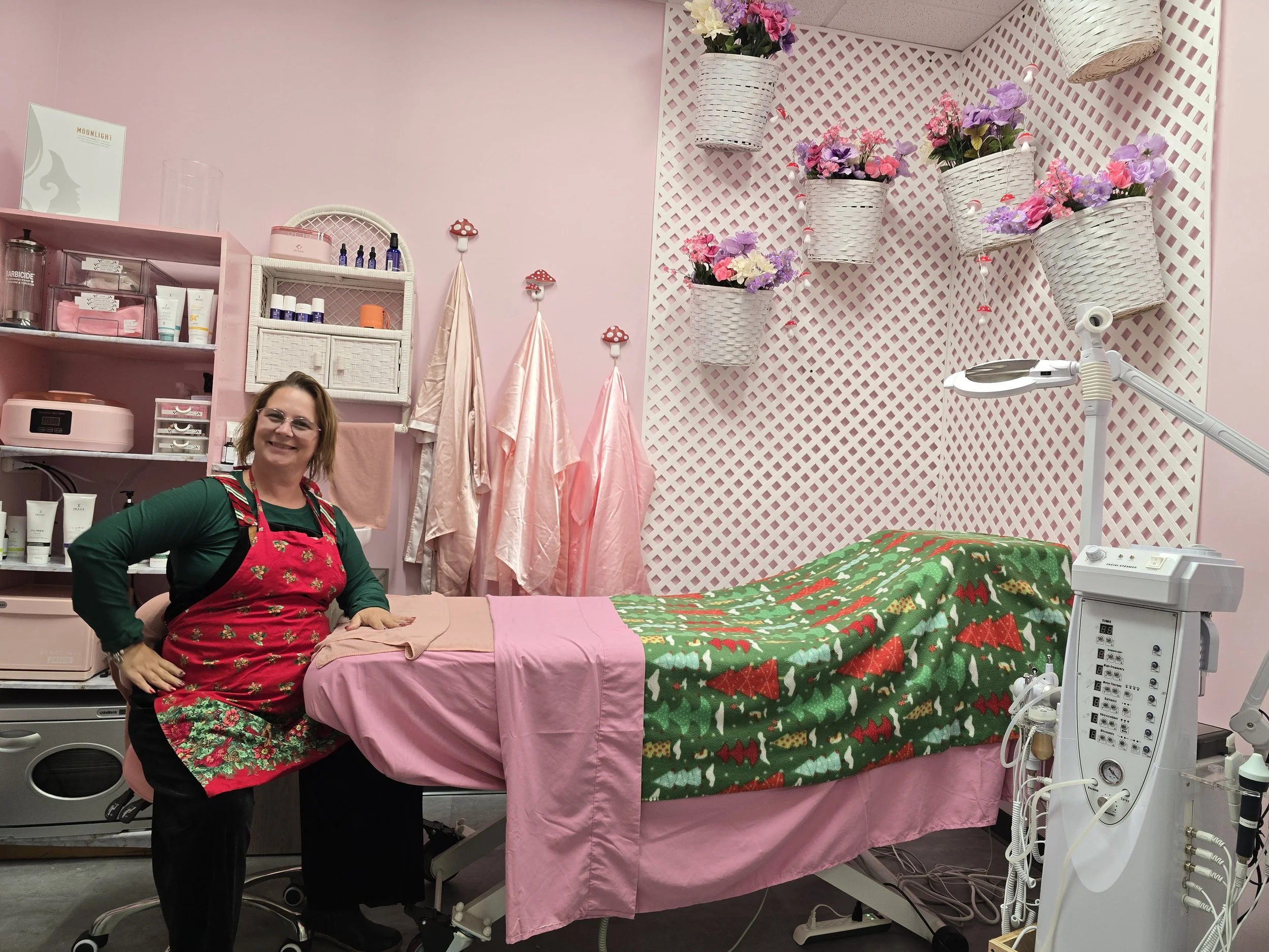 A woman in an apron smiling next to a facial table in a pink facial studio decorated with flower baskets and white lattice wall panels.
