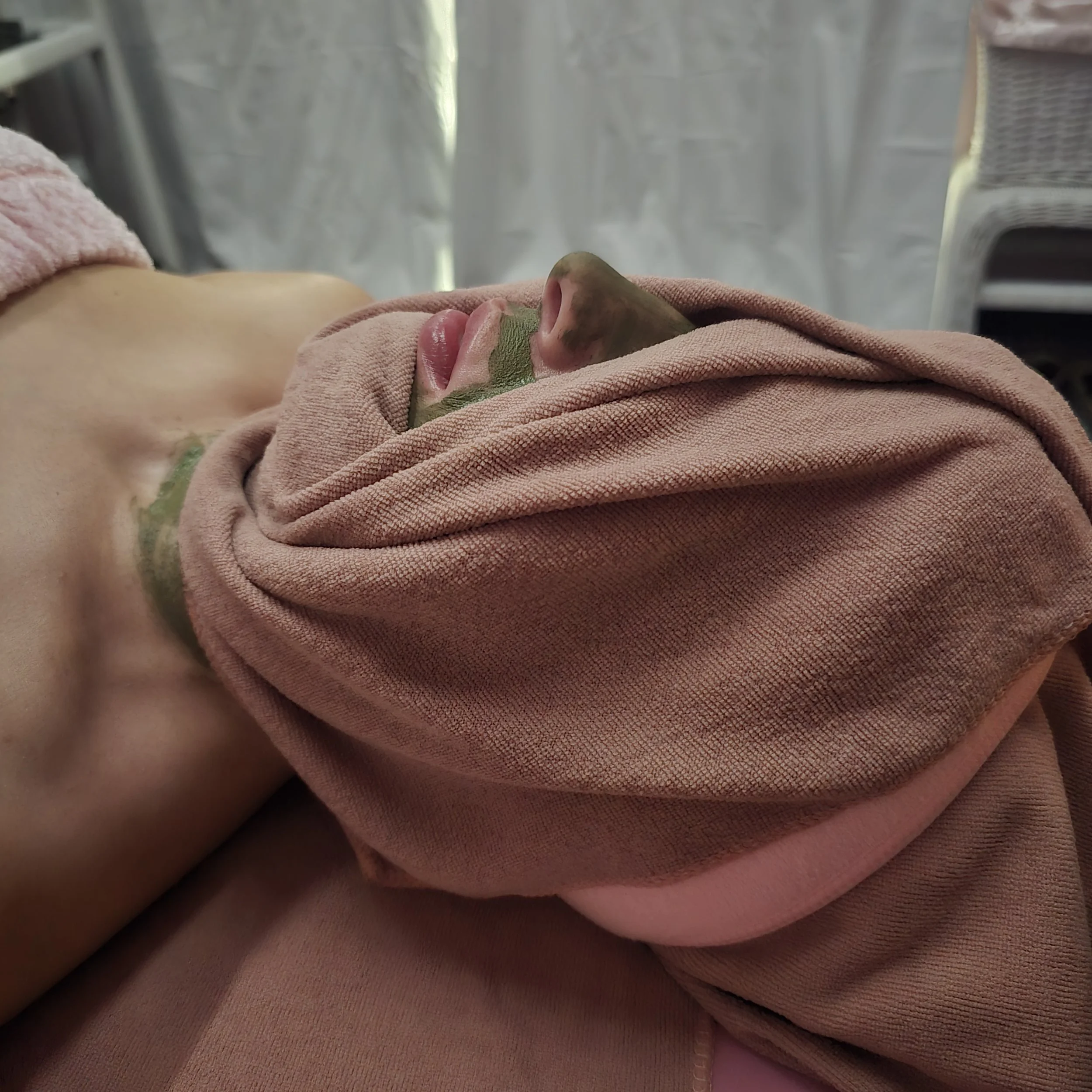 A person lying down with their face partially covered by a pink fabric while receiving a green facial mask, with indoor curtains and furniture in the background.