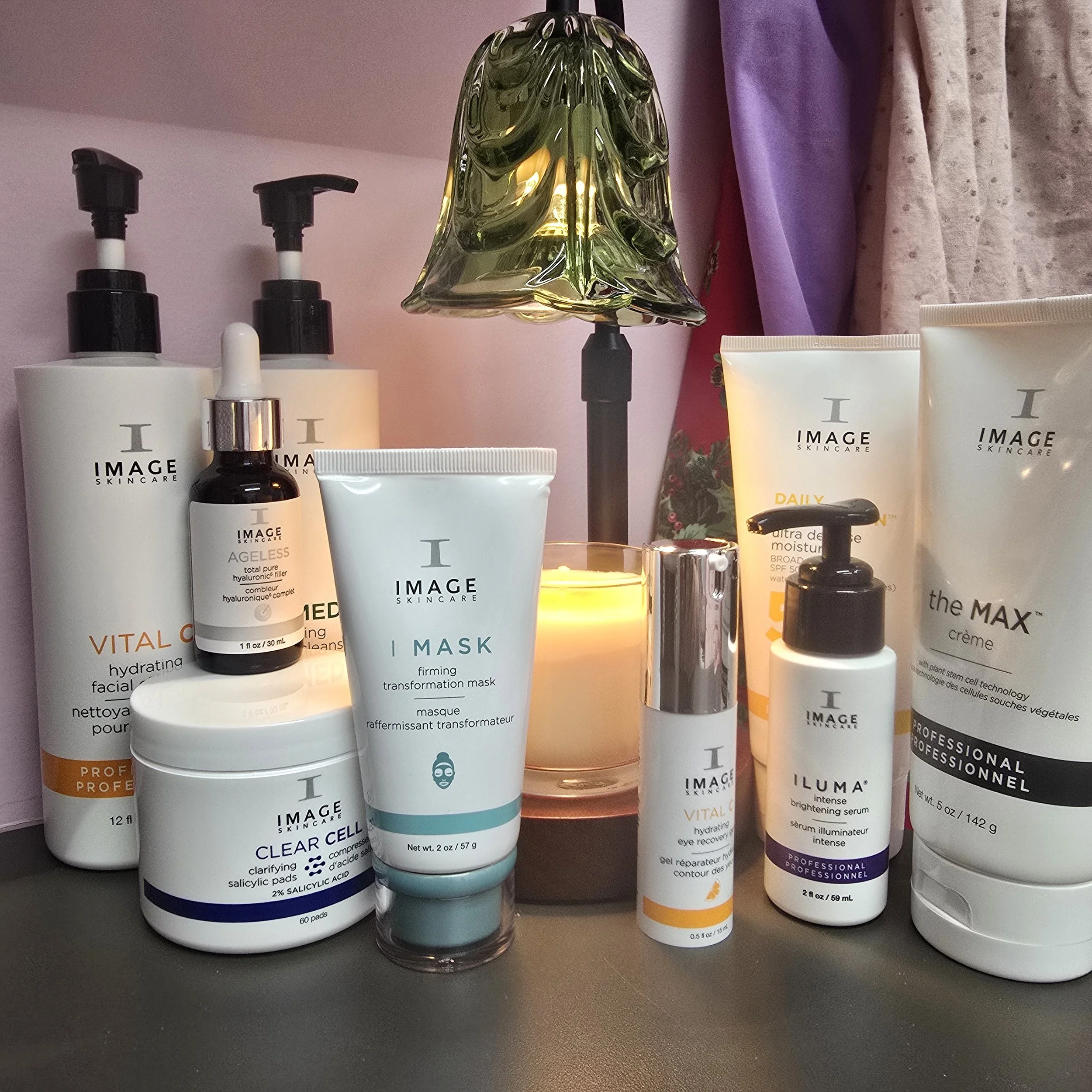 Collection of skincare products on a nightstand with a small lamp, candles, and decorative curtains in the background.