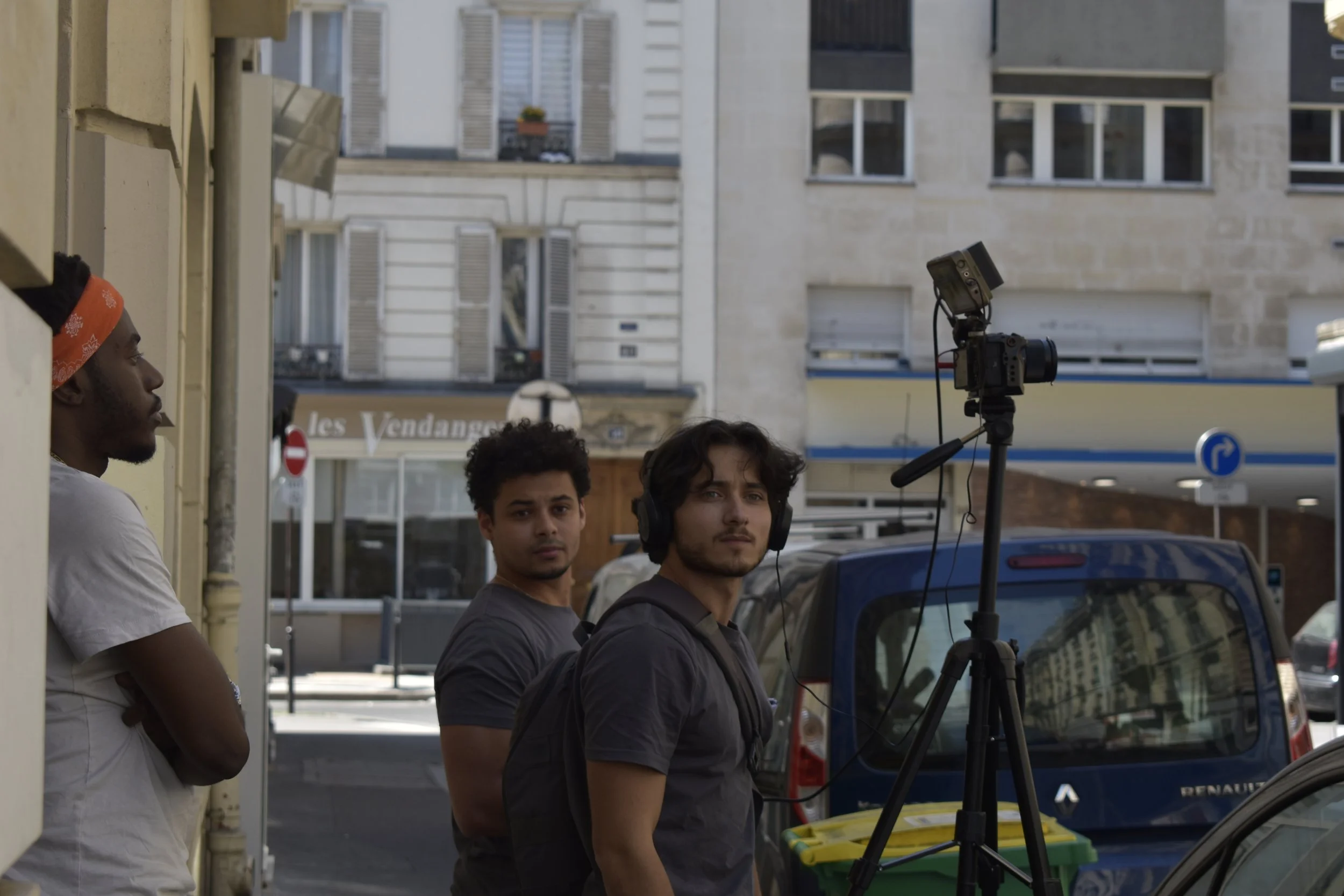 Trois jeunes hommes se tiennent sur un trottoir dans une rue urbaine, avec un appareil de tournage fixé sur un trépied. L'un porte un casque d'écoute et regarde attentivement, un autre porte un bandeau orange, et le troisième regarde directement la caméra.