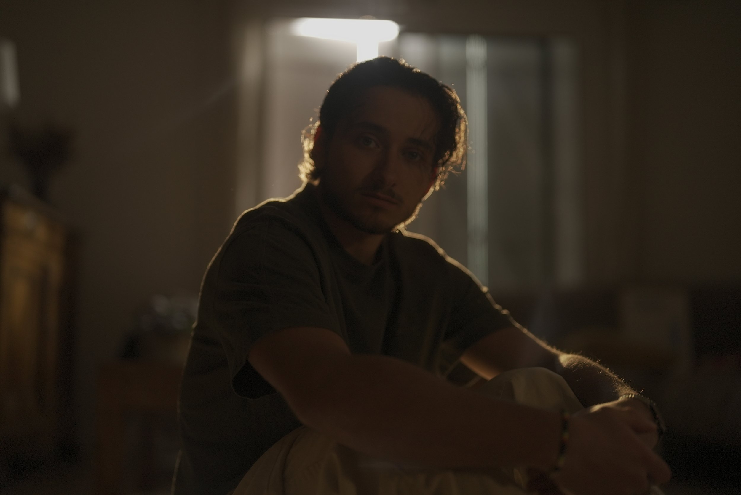 Homme assis dans une pièce sombre avec lumière venant d'une fenêtre derrière lui, expression pensive.