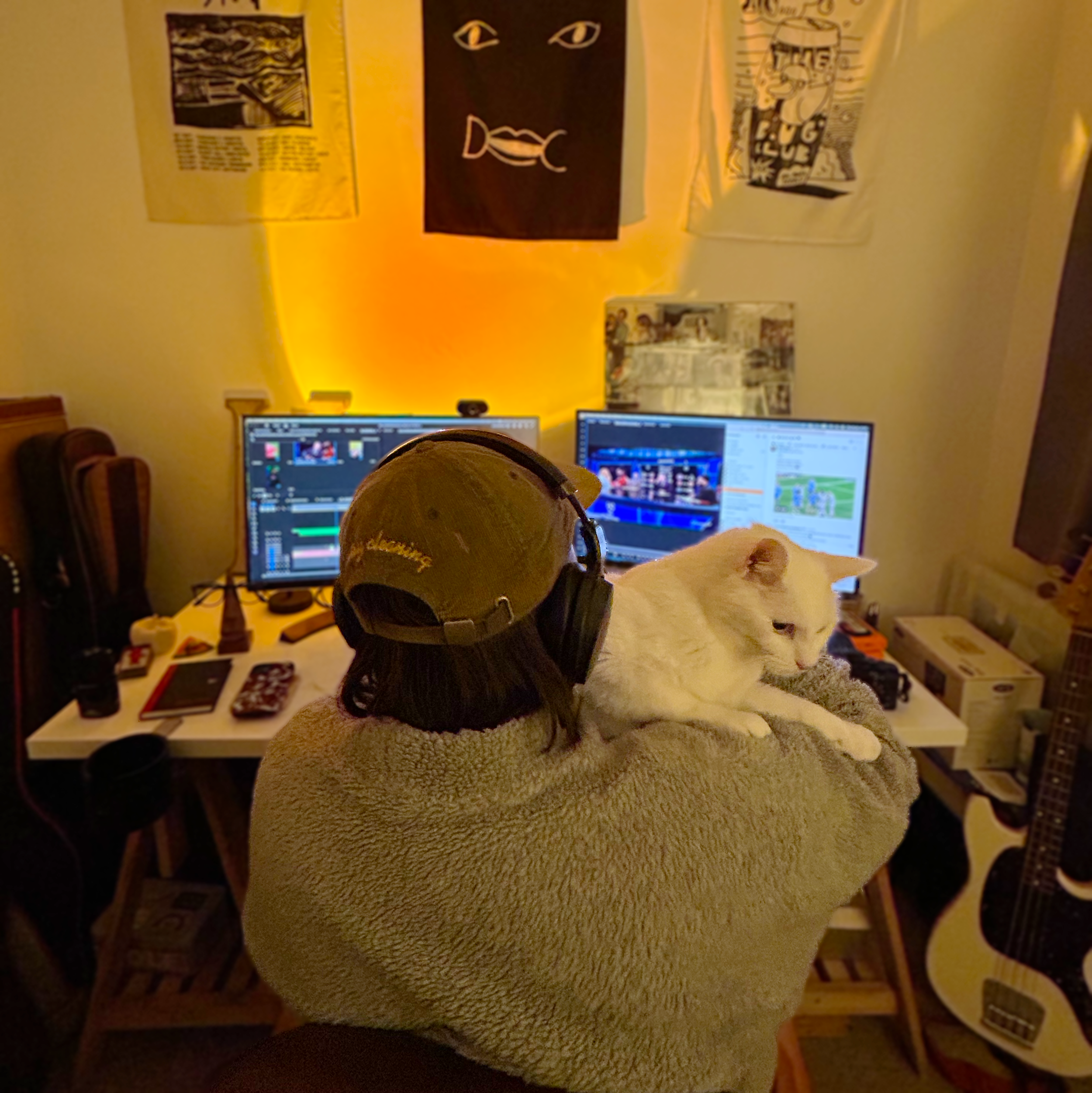 Still image of Harvey sat at his editing desk, holding a white cat. The cat faces the camera and Harvey faces away looking at two landscape slightly blurred computer screens.