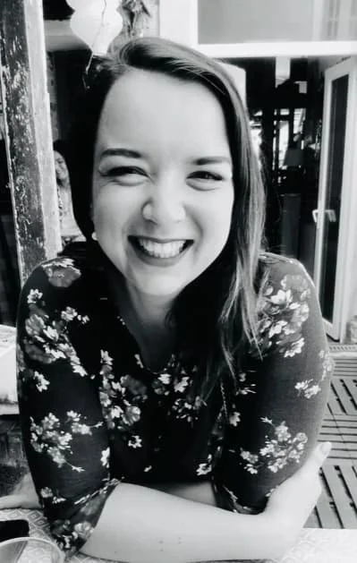 Une femme souriante avec une robe à fleurs, assise dans un espace intérieur, en noir et blanc.