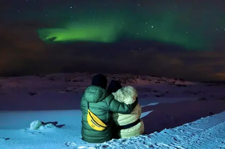 Deux personnes de dos, assises dans la neige, regardant une aurore boréale dans le ciel islandais