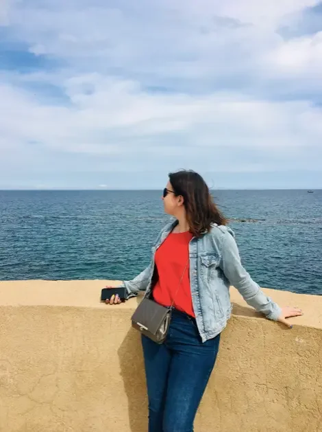 Femme debout près d'une balustrade en vue de l'océan, portant un jean, un débardeur rouge, une veste en jean noire et des lunettes de soleil, tenant un téléphone portable