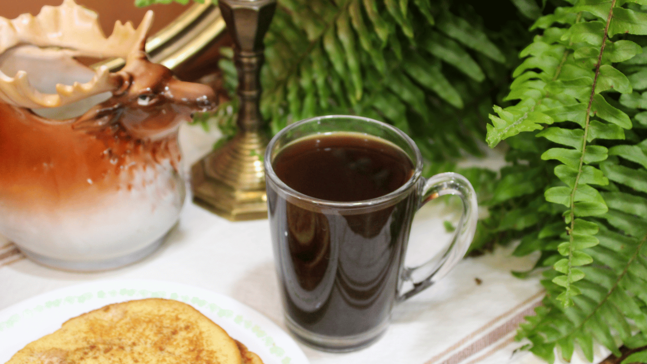 A breakfast setup with a plate of French toast, a mug of black REPOTTED coffee, a decorative ceramic pot, a brass candlestick, and green ferns in the background.