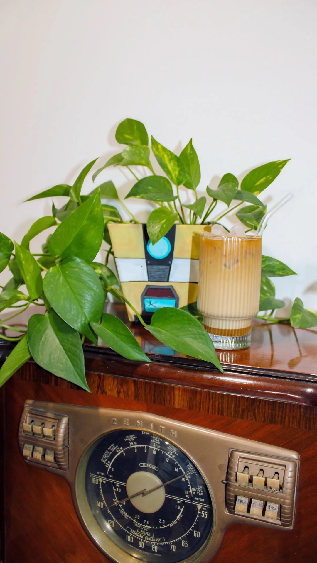 A vintage radio with a plant in a colorful pot and a glass of iced coffee with a spoon on a wooden surface.