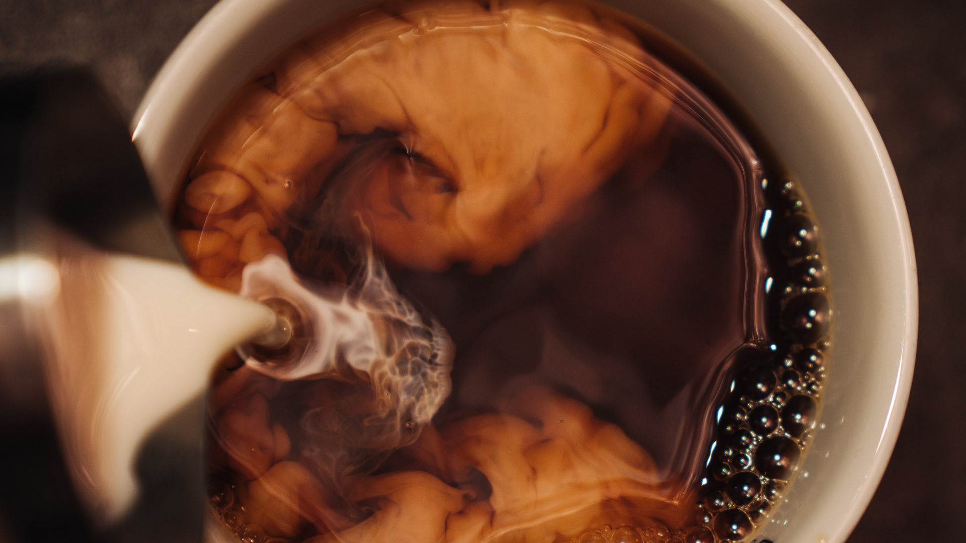 Hot coffee with cream being poured into a white mug, creating swirling patterns in the dark liquid.