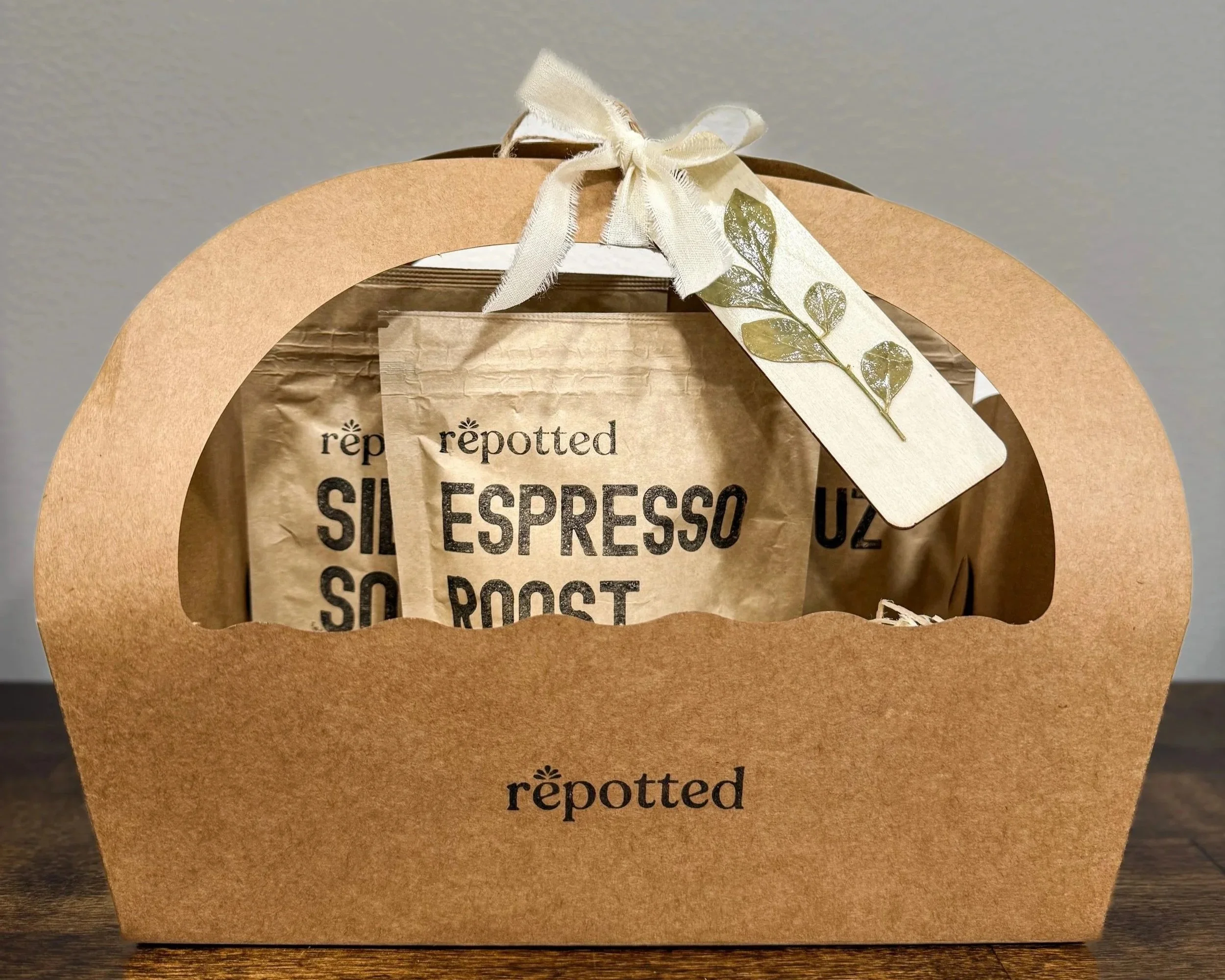A brown paper gift bag with a clear window revealing repotted coffee bags inside, tied with a white ribbon and decorated with a white tag featuring a preserved green leaf.
