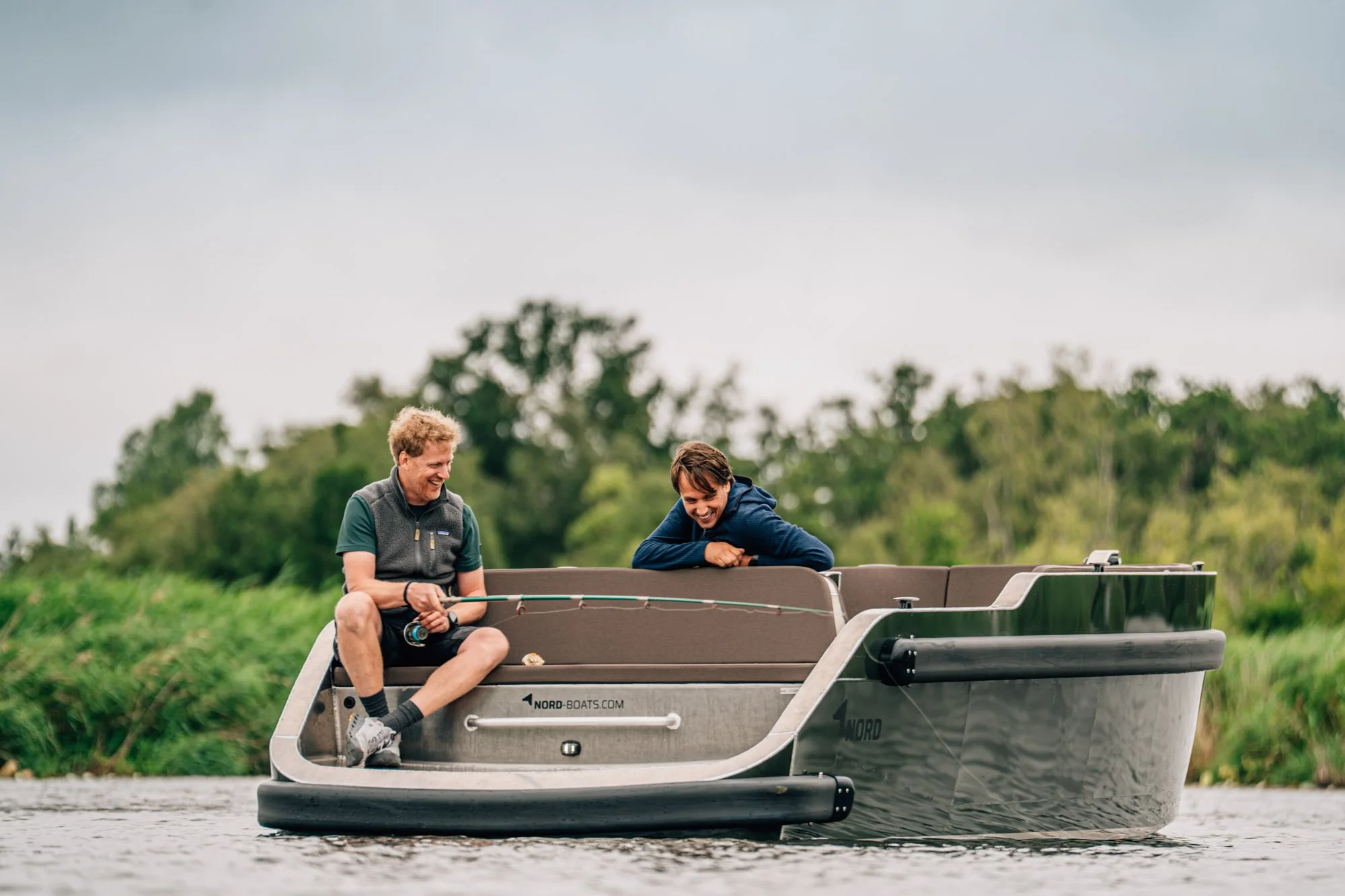 Twee jonge mannen lachen op een grijs bootje in een rivier, met een achtergrond van groen landschap en een bewolkte lucht.