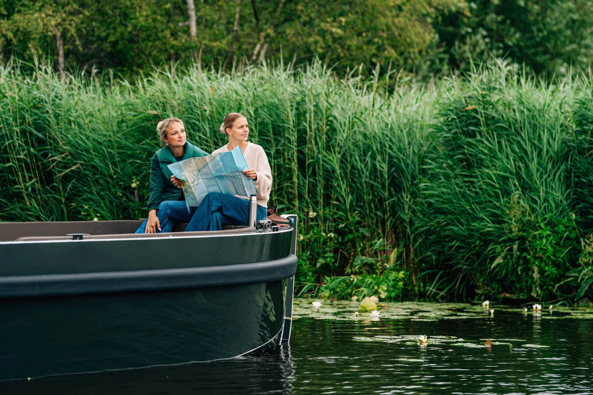 Twee vrouwen zitten in een boot op een meer, met een grote kaart in hun handen, omgeven door groene riet en bomen.