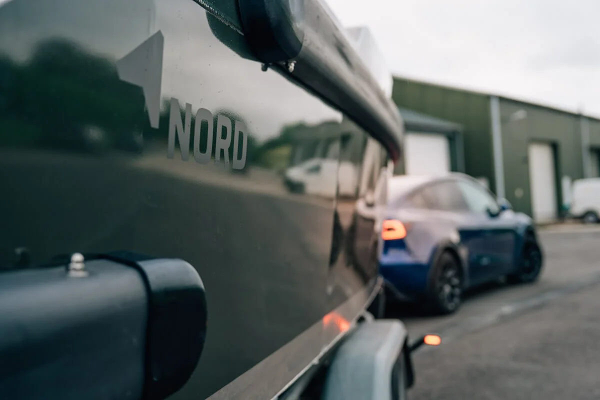 Close-up van een voertuig met de tekst 'NORD' en een pijl op de zijkant, met een sportauto op de achtergrond in een industriële omgeving.