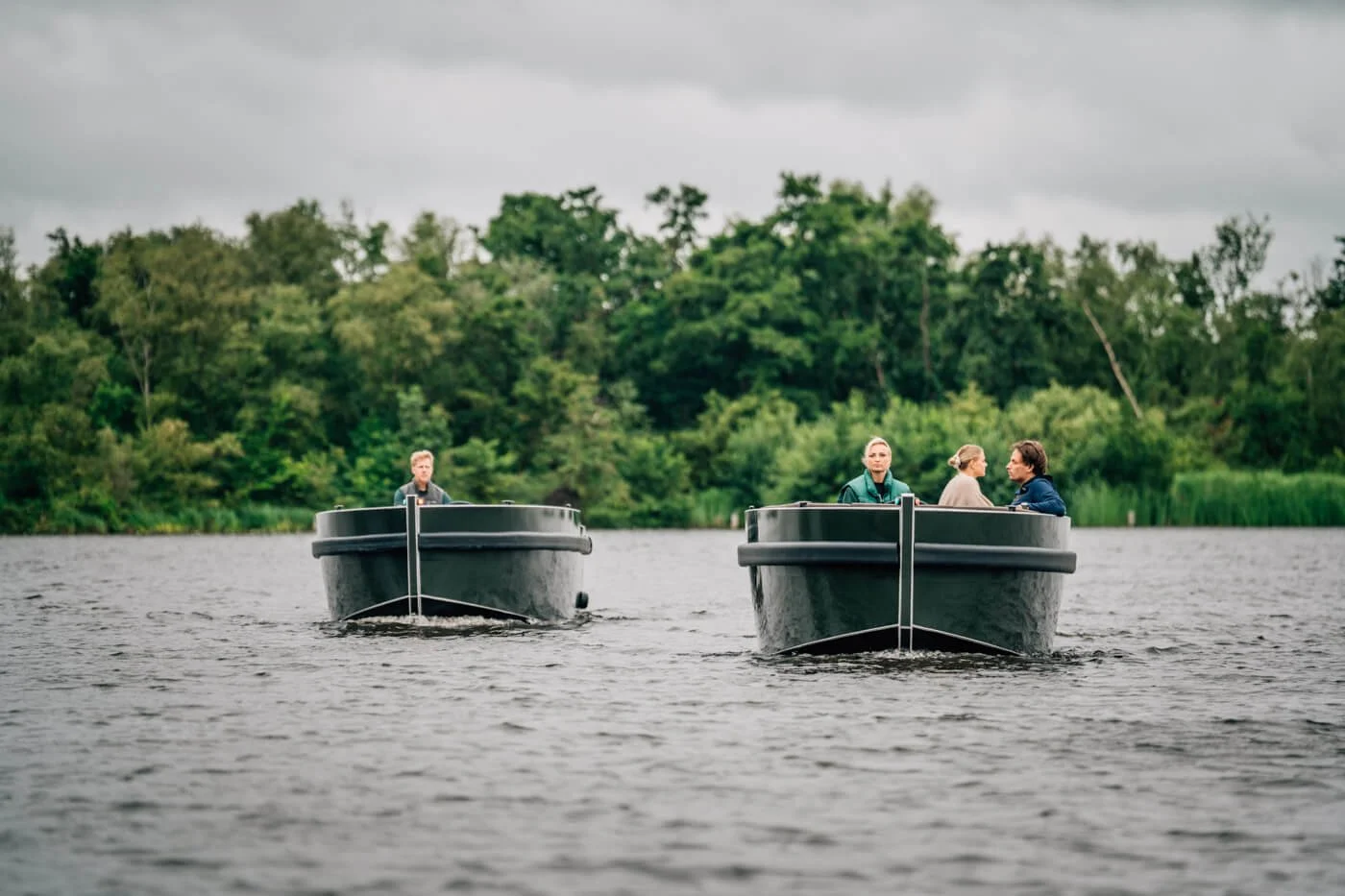 Twee boten op een kalme rivier met drie personen aan boord, achtergrond van groene bomen onder een bewolkte hemel.