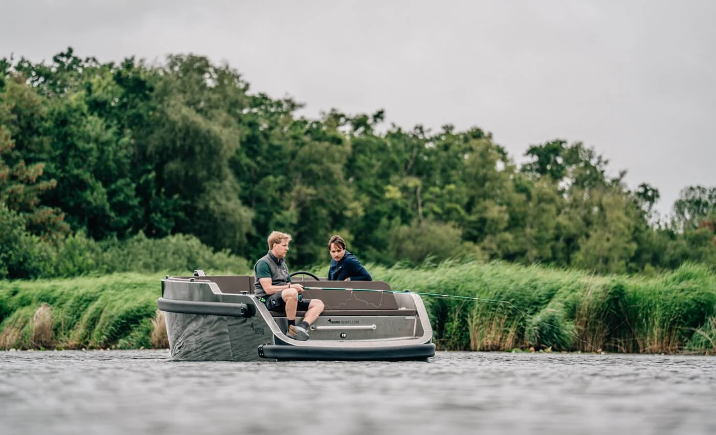 Twee mensen vissen op een kleine motorboot op een rivier, omringd door groene bomen en riet.