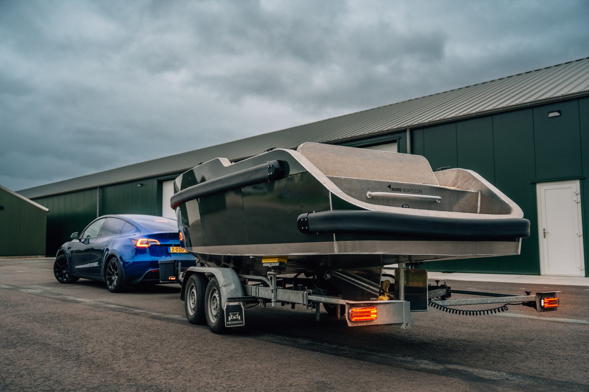 Een blauwe Tesla-auto trekt een bootwagon met een grote boot op een parkeerplaats, naast een groen industrieel gebouw onder een bewolkte hemel.