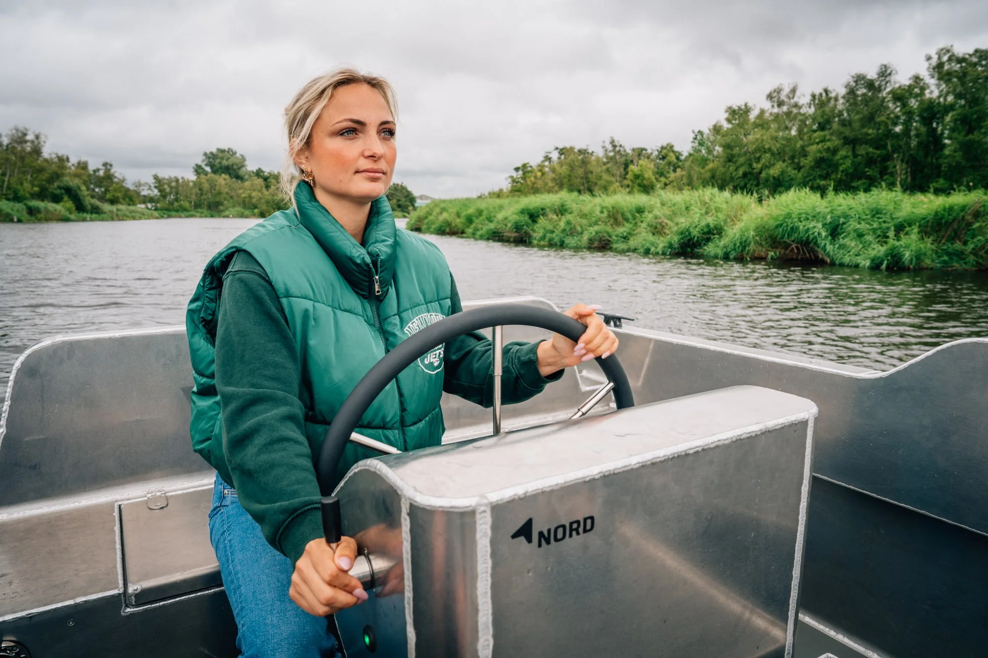 Vrouw die een boot bestuurt op een rivier, omgeven door groen landschap en bewolkte hemel.