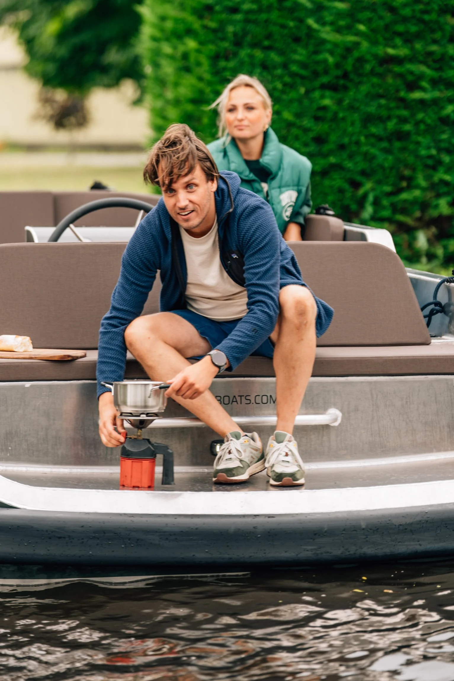 Een man in sportkleding die een gasfles onder een boot bevestigt, terwijl een vrouw op de achtergrond in een groene jas naar hem kijkt.