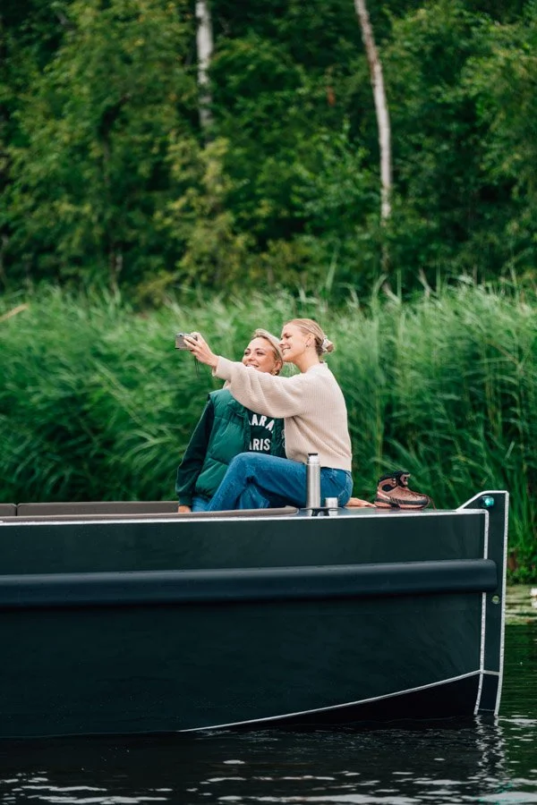 Twee vrouwen maken een selfie op een boot, met groen bos op de achtergrond.