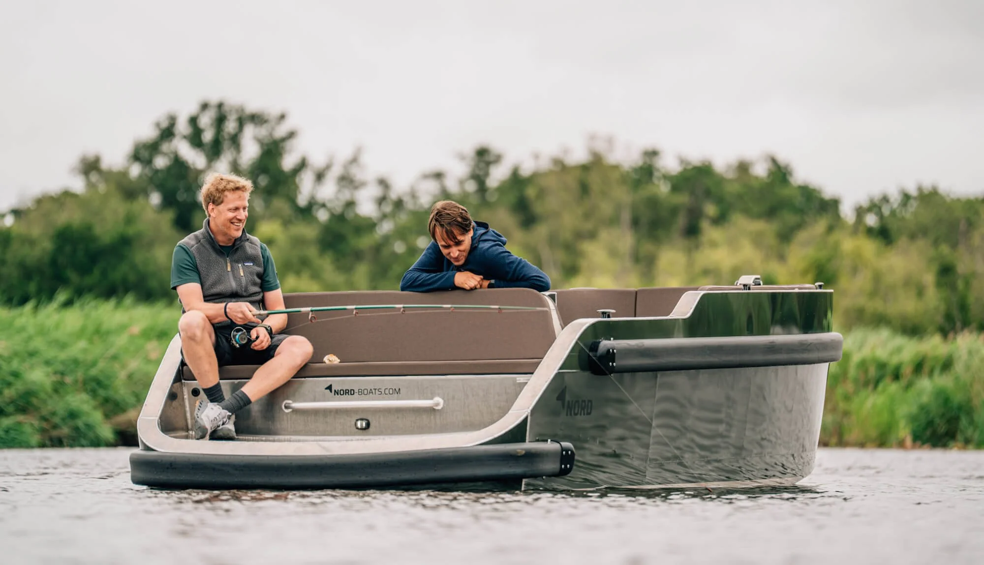 Twee mannen zitten op een boot, de ene is aan het vissen en de andere lacht.
