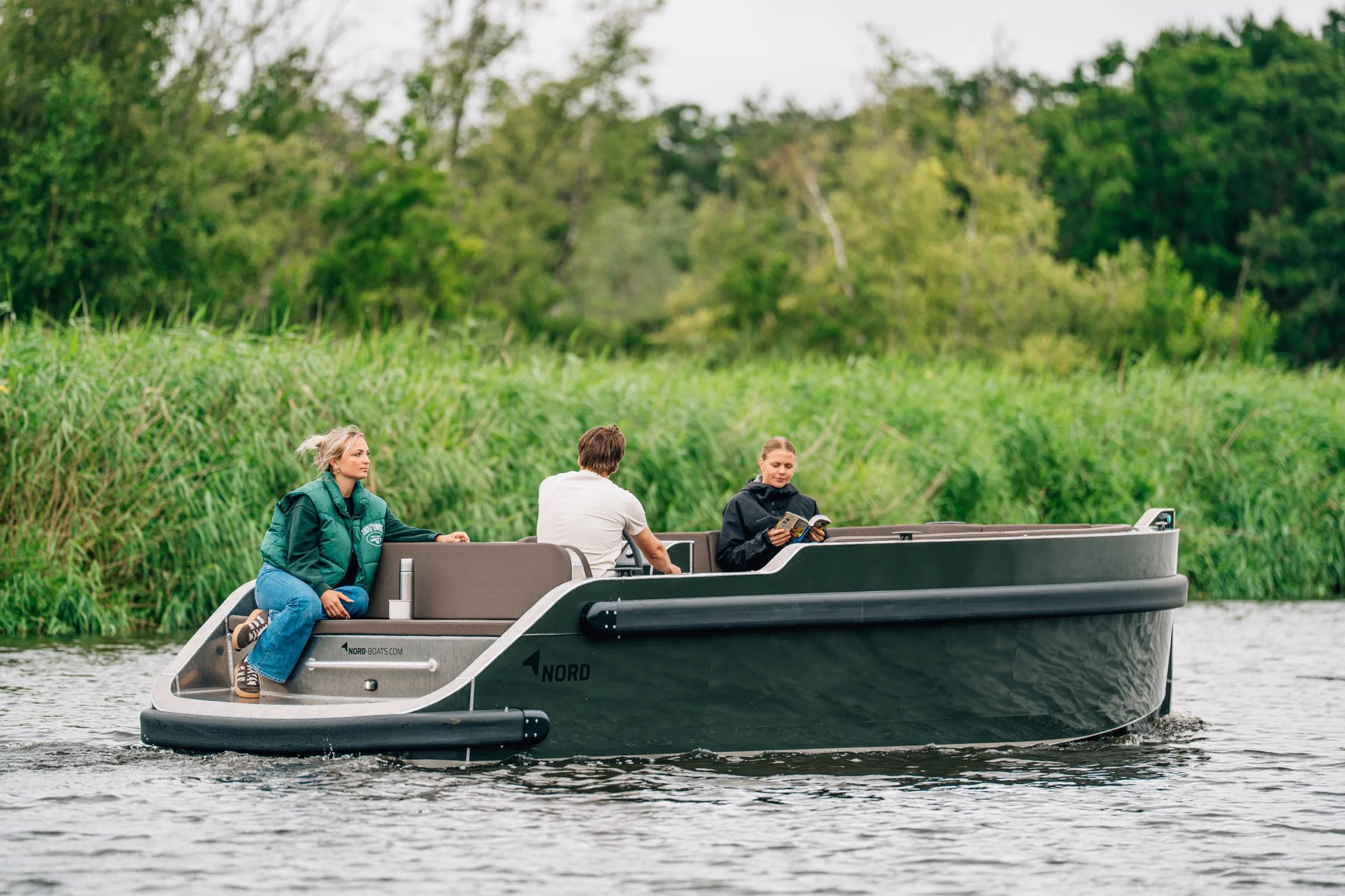 Drie mensen op een elektrische boot op een rivier met groene struiken en bomen op de achtergrond, twee vrouwen en een man, een vrouw leest een tijdschrift.