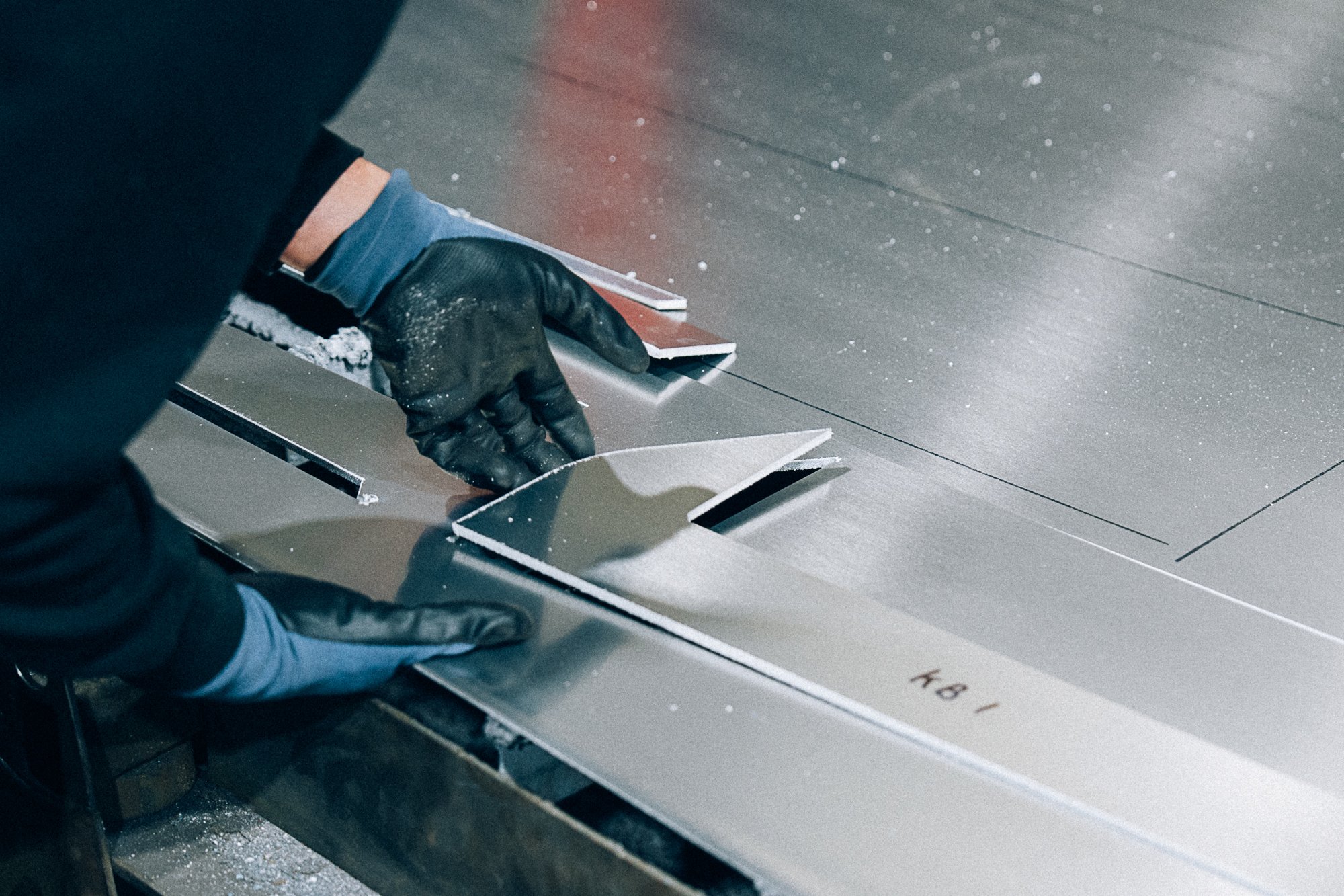 Persoon die metalen plaat installeert met behulp van een spatel, iemand draagt zwarte handschoenen, de plaat ligt op een werkbank, en het oppervlak is stoffig met kleine deeltjes en vuil.