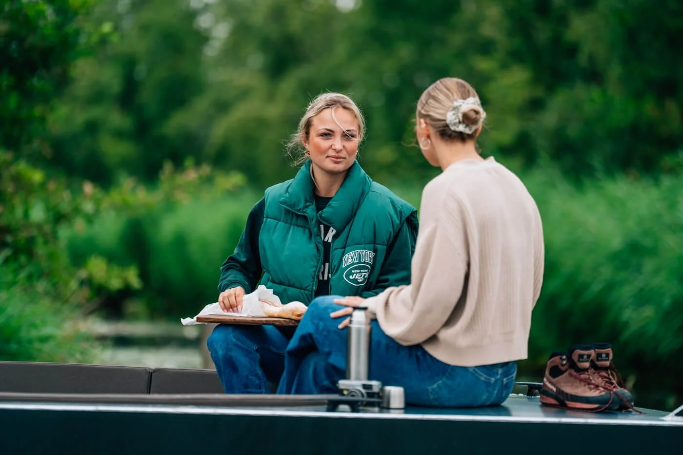 Twee vrouwen zitten op een boot in een groene omgeving, ze praten met elkaar en een van hen houdt een bord eten vast.