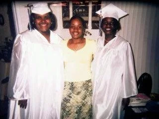 Two women in graduation gowns and caps standing with a woman in a yellow top and patterned skirt in front of a window.