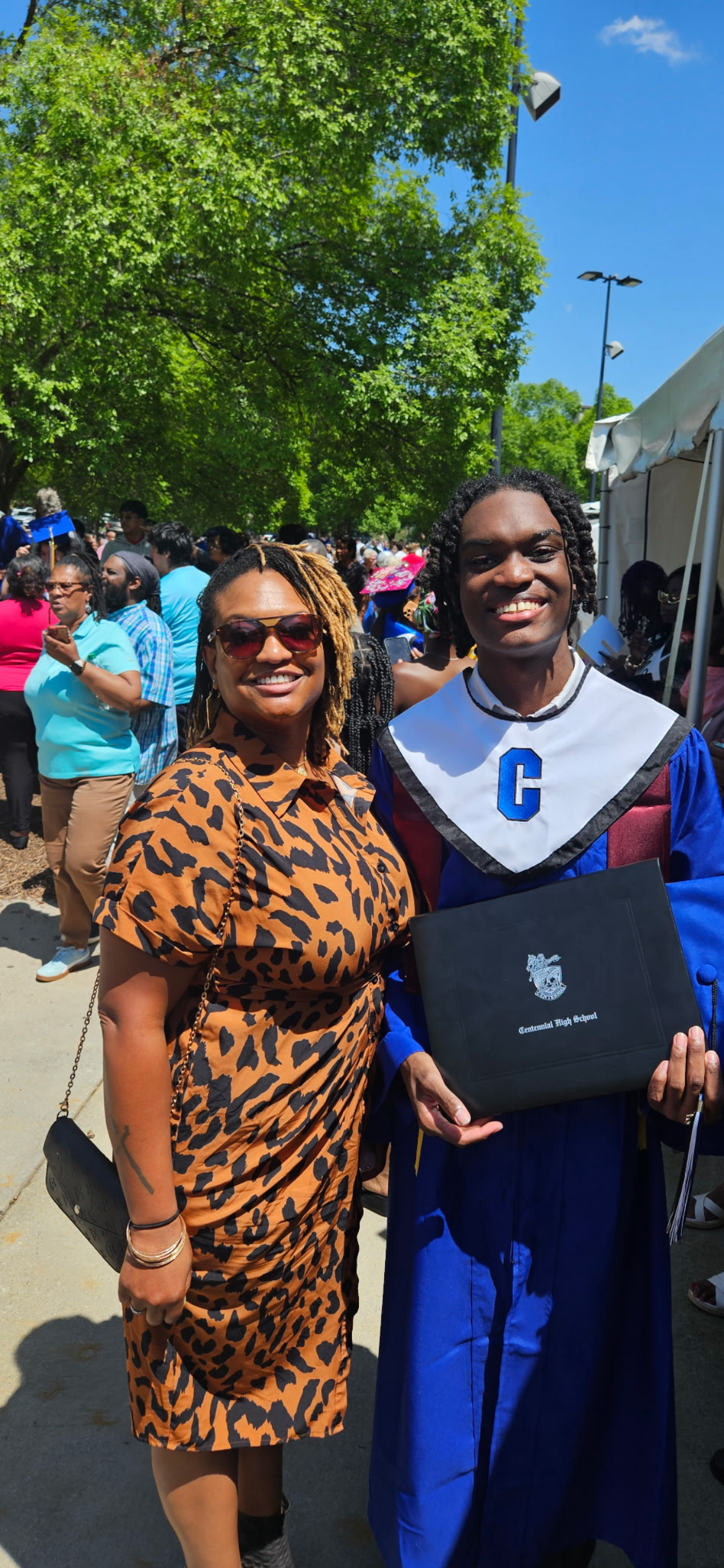 A young man in a blue graduation gown and cap holding a high school diploma, standing next to his mother in a leopard print dress, both smiling at an outdoor graduation ceremony with many people and green trees in the background.