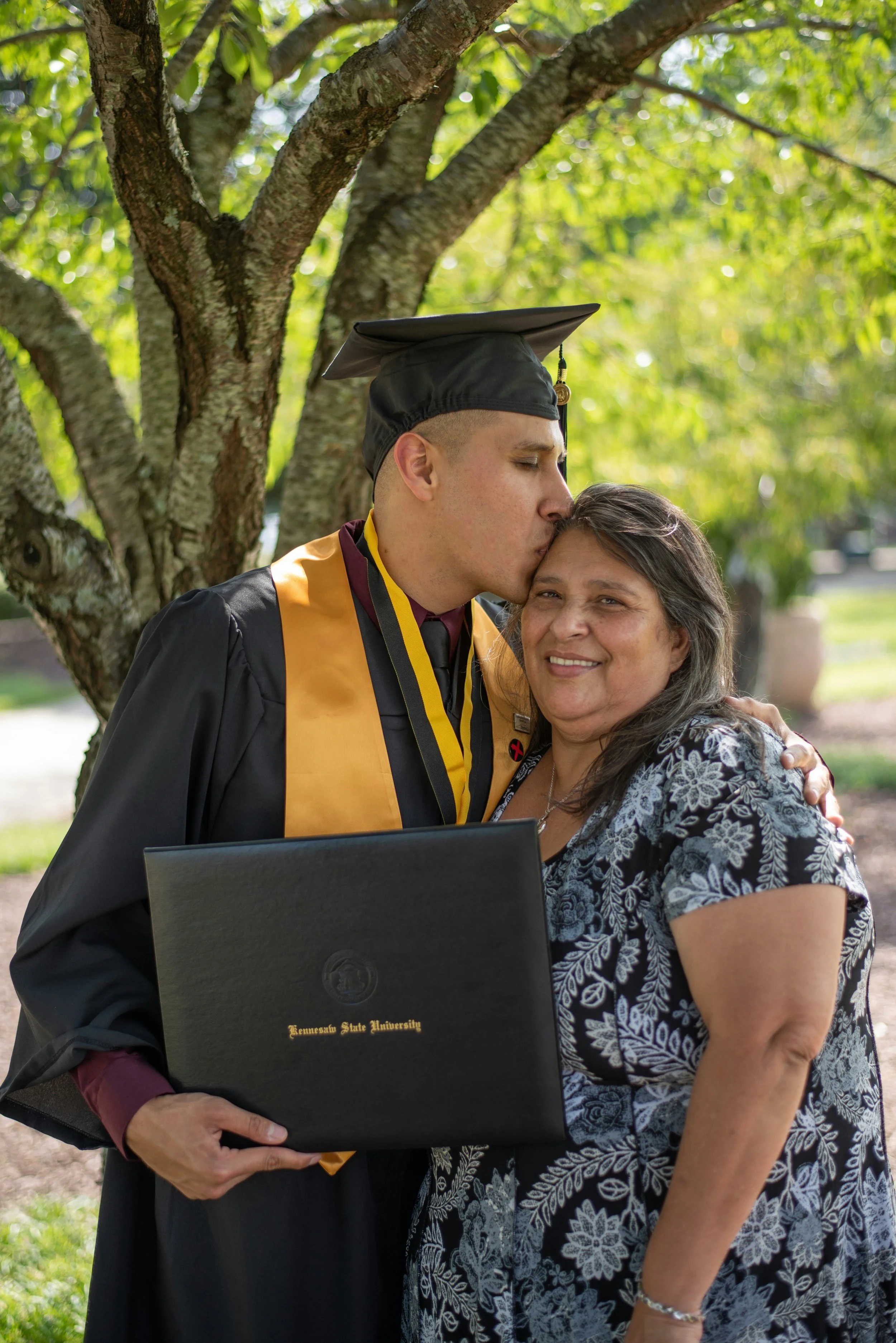 A graduate in a black cap and gown with a gold and black stole kisses his smiling mother on the forehead. The graduate holds a high school diploma in his left hand. They are outdoors under a tree with green leaves.