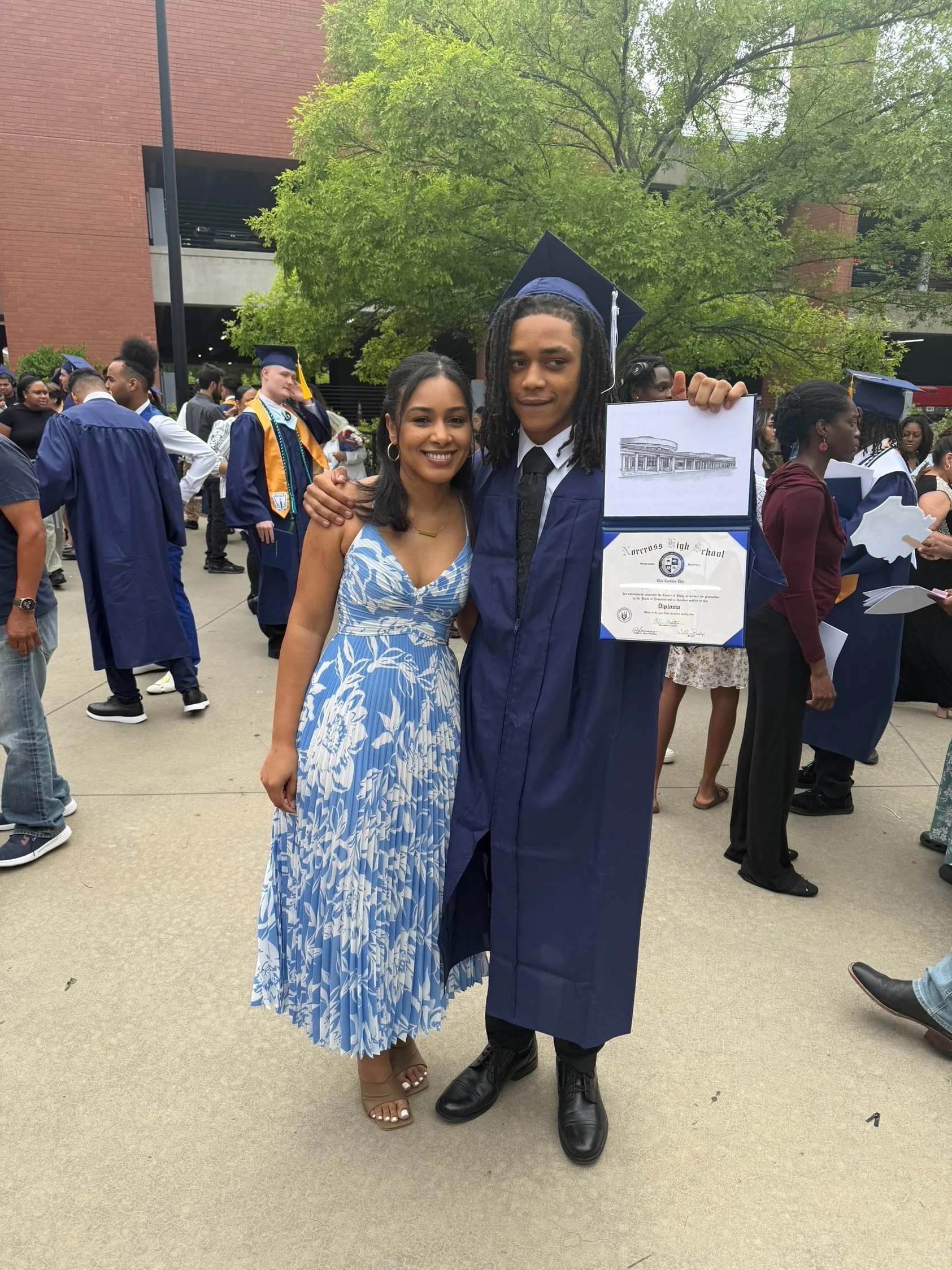 A young man in a navy high school graduation gown, cap, and tie holding his diploma, smiling and standing next to a woman in a blue and white dress. They are outdoors during graduation celebration with other graduates and people in the background.