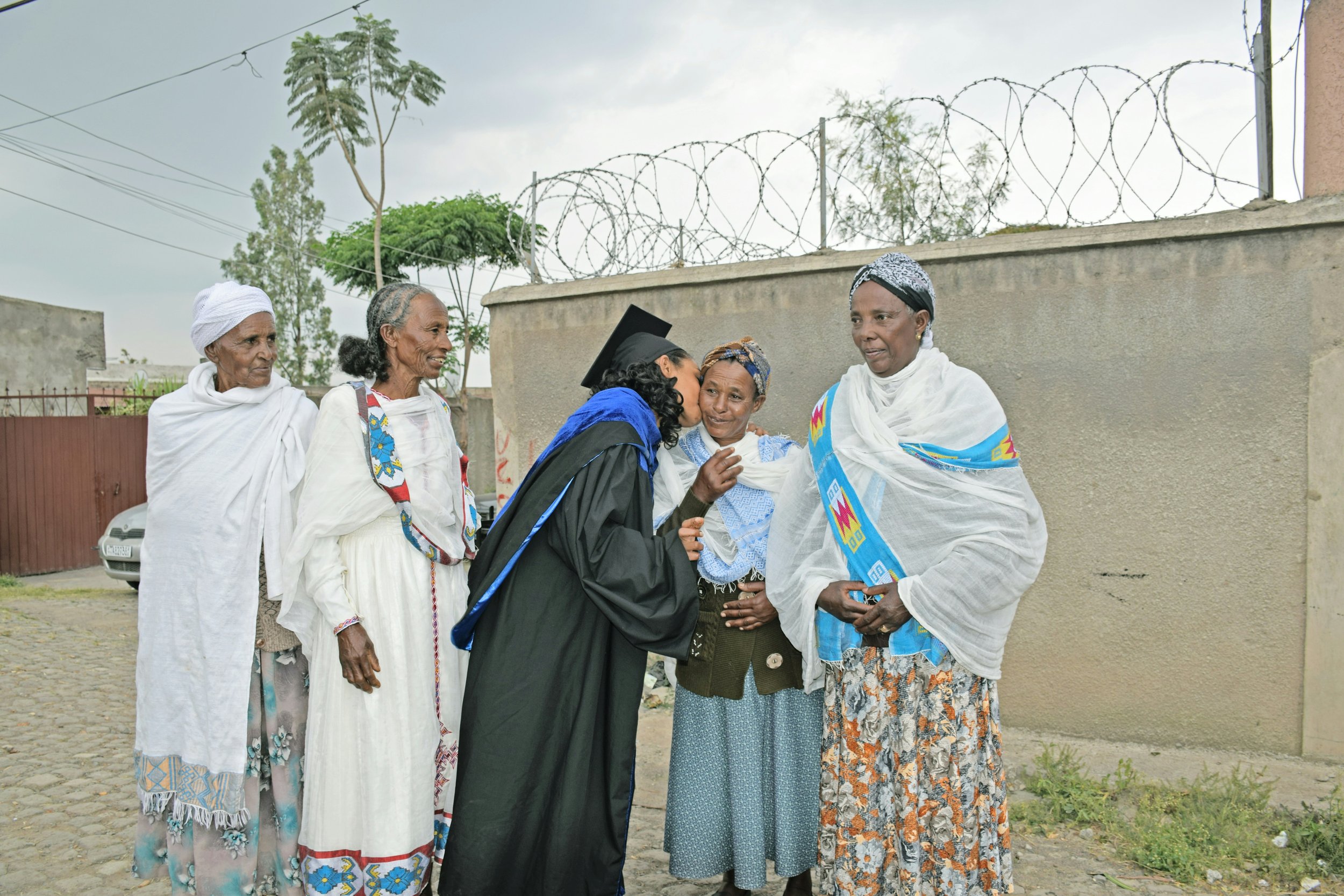 A young woman in a graduation gown and cap is kissing an elderly woman on the cheek. The women are wearing traditional African clothing and headscarves.