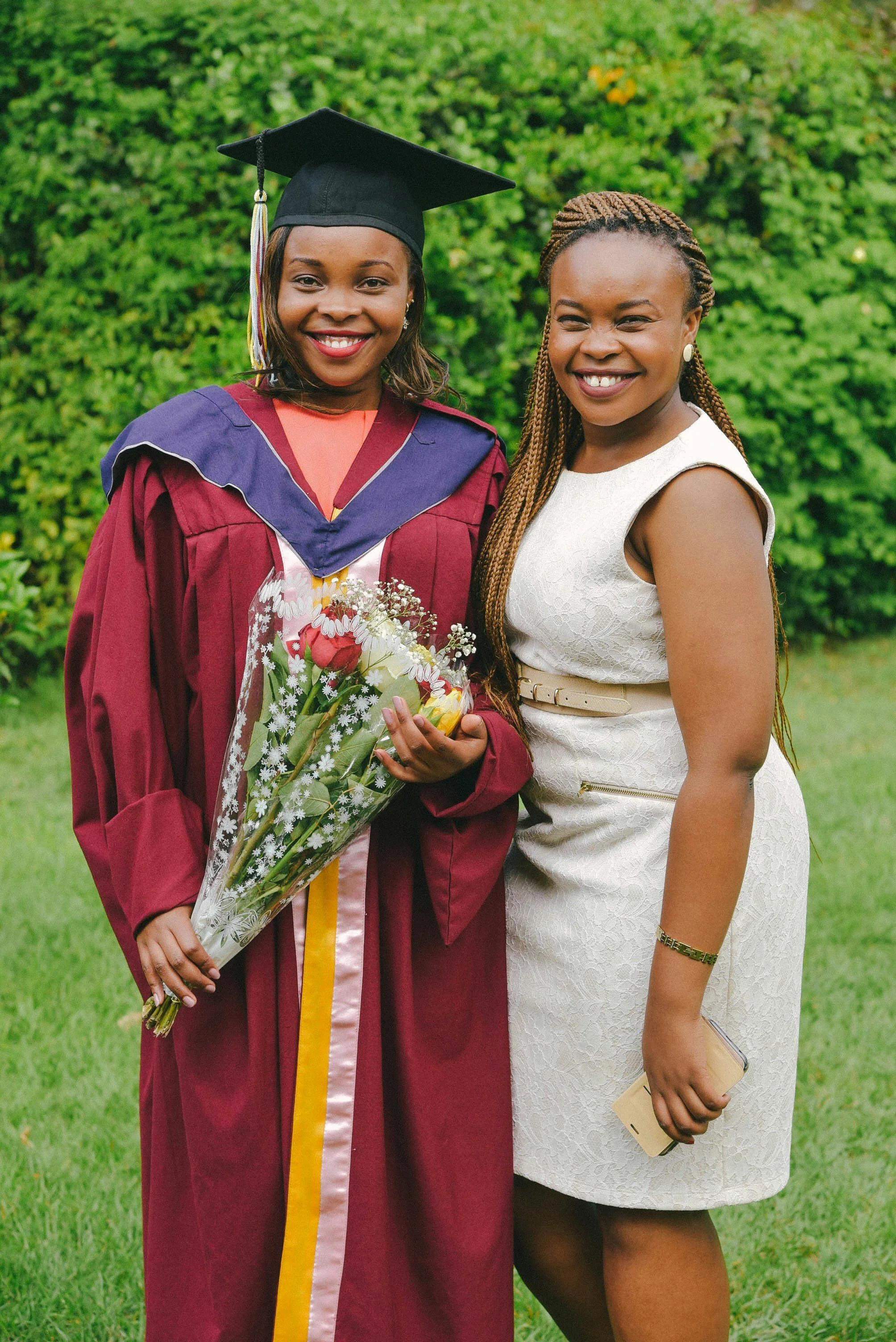 A high school girl in graduation gown and cap holding a bouquet of flowers standing next to her mother in a white dress outdoors.