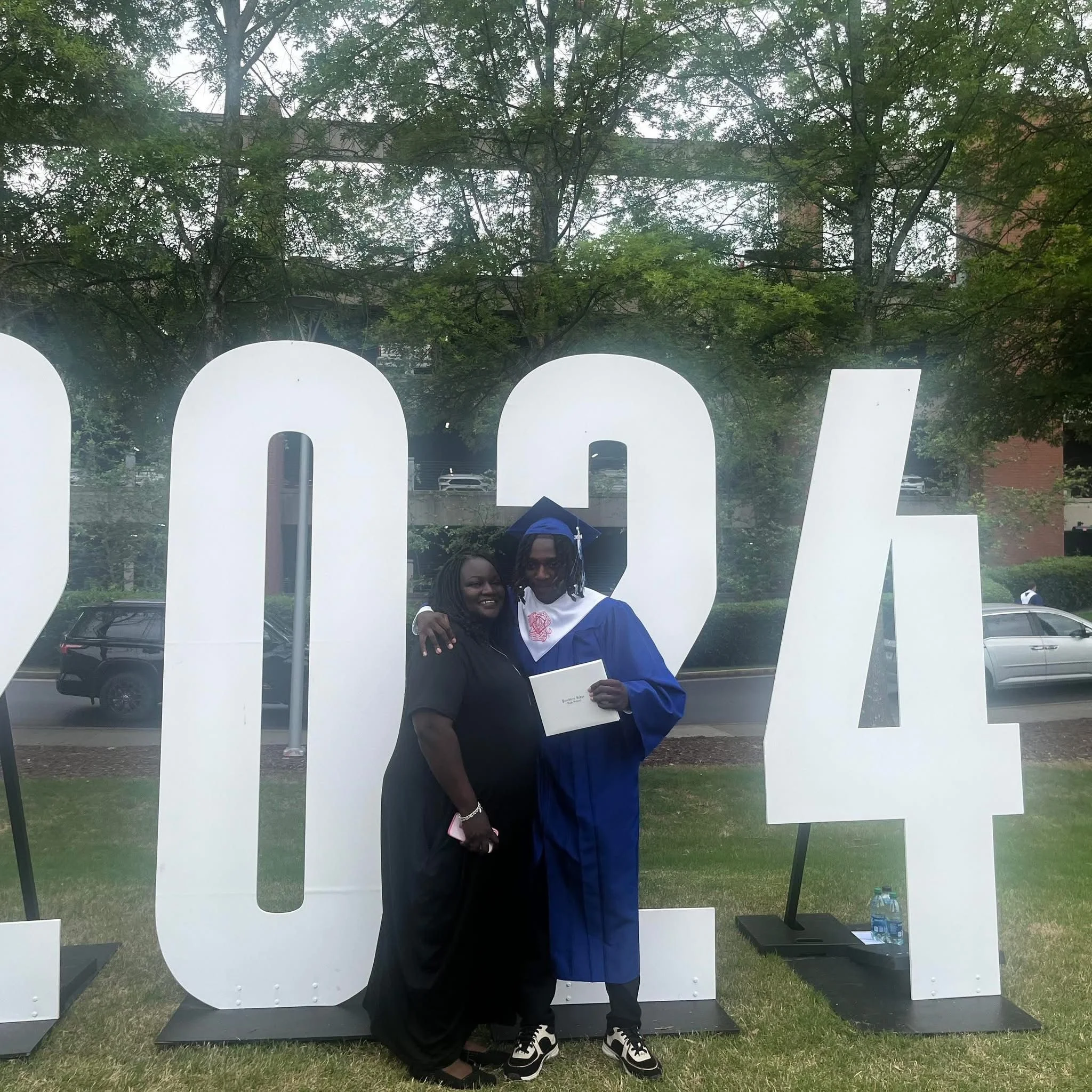 A graduate in blue cap and gown holding a high school diploma, standing next to his mother in black dress, both smiling, with large white 2024 sign and trees in the background.