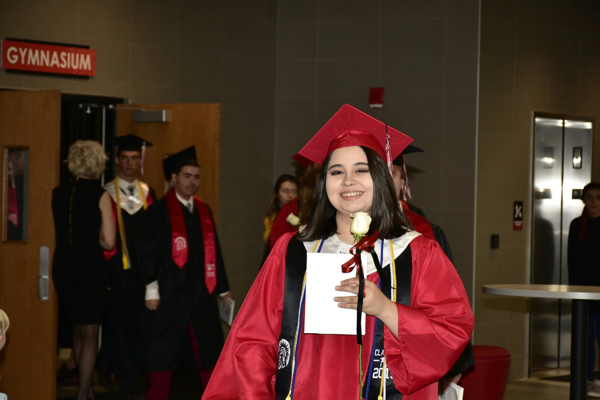 Young woman in red high school graduation gown and cap holding diploma and white rose, smiling at graduation ceremony.