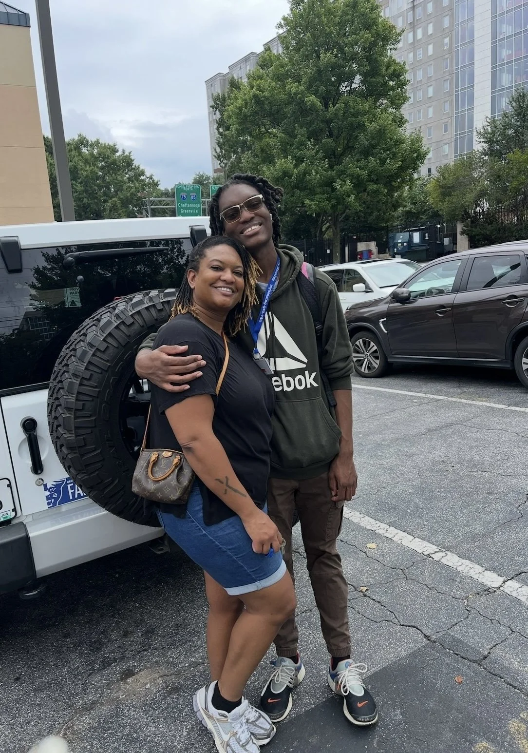 A single mother and her son, a senior in high school, smiling and hugging each other in a parking lot, with a white vehicle behind them and trees and buildings in the background.