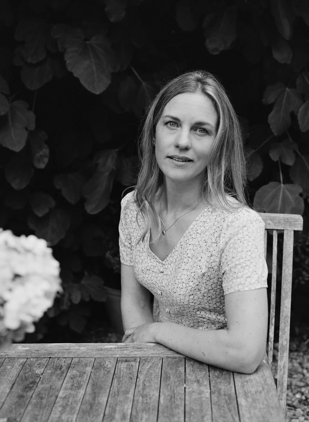 A woman with light-colored hair and a light-colored, patterned short-sleeve shirt sits at a wooden table outdoors against a background of large leaves.