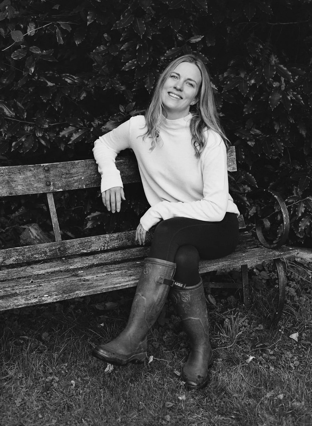 A woman sitting on a wooden bench outdoors, wearing a white sweater, dark leggings, and rubber boots, with lush foliage in the background.