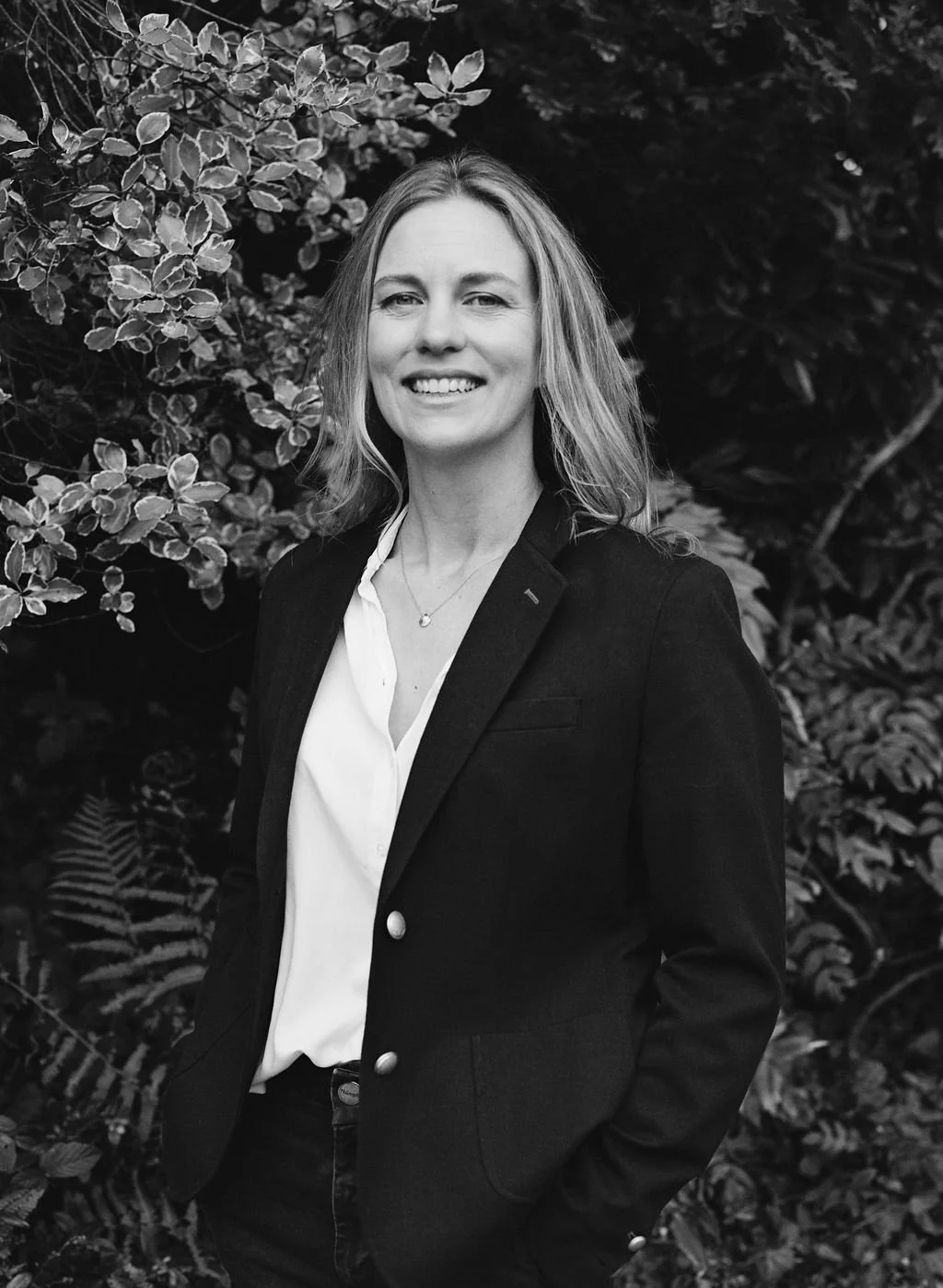 A woman smiling in front of leafy bushes in black and white.
