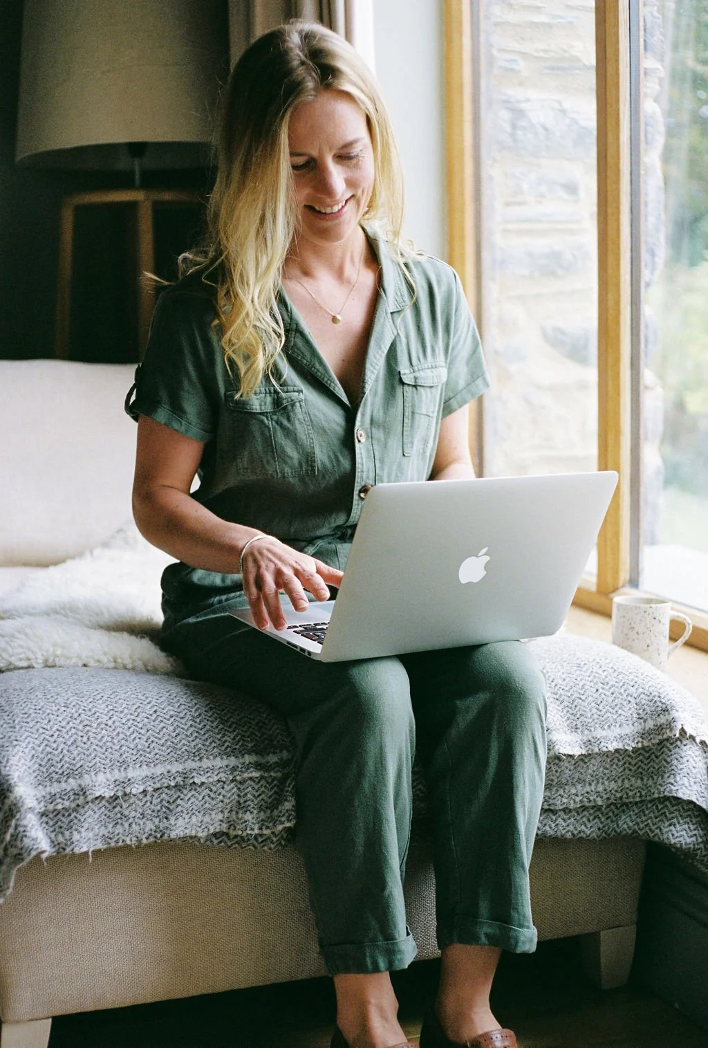 A woman with blonde hair smiling while using a silver MacBook laptop, sitting on a cozy window seat with a white blanket, near a large window with a view of greenery outside.