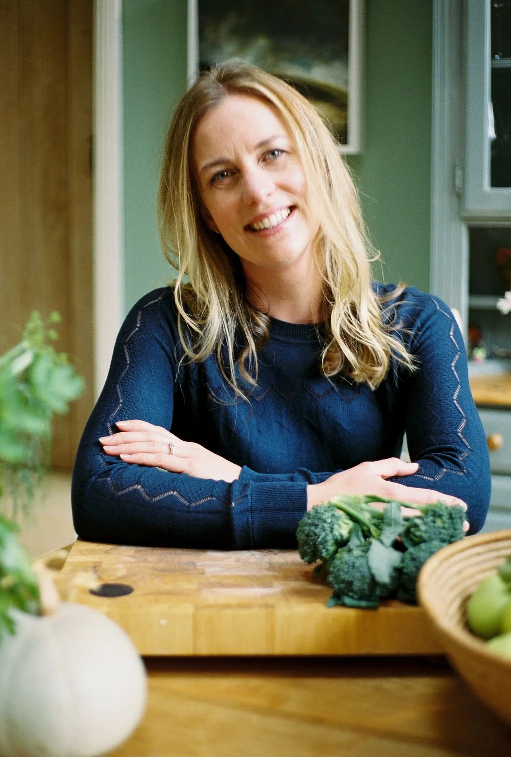 Rosamund Yoxall, smiling and sitting at a wooden table in a kitchen, with broccoli and other vegetables in front of her.