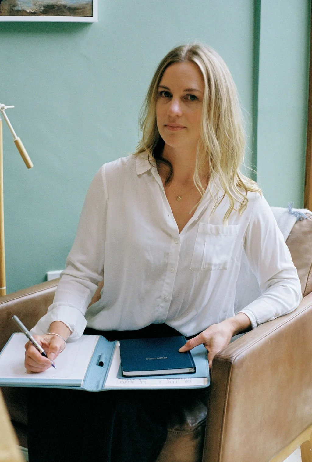 UK Health Strategist sitting on a brown leather chair, holding a pen and writing in an open notebook on her lap, with a green wall and framed picture in the background.