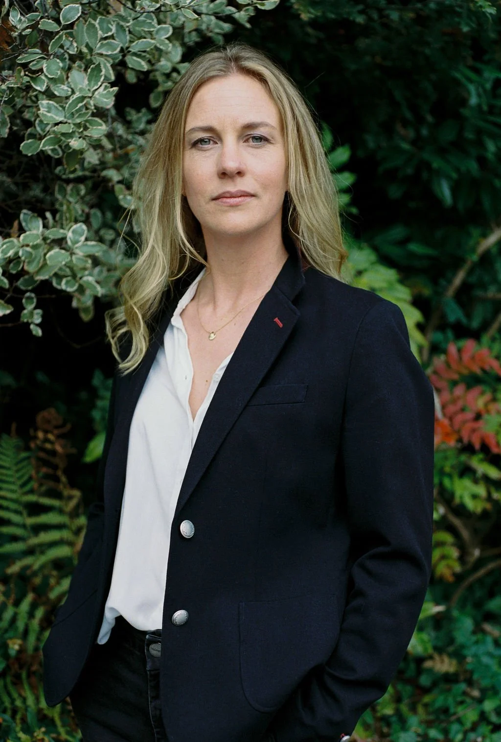 Head shot of Dr Rosamund Yoxall, Health Strategist in the UK, writer, consultant and speaker, in a black blazer and white shirt standing outdoors in front of green leafy plants.