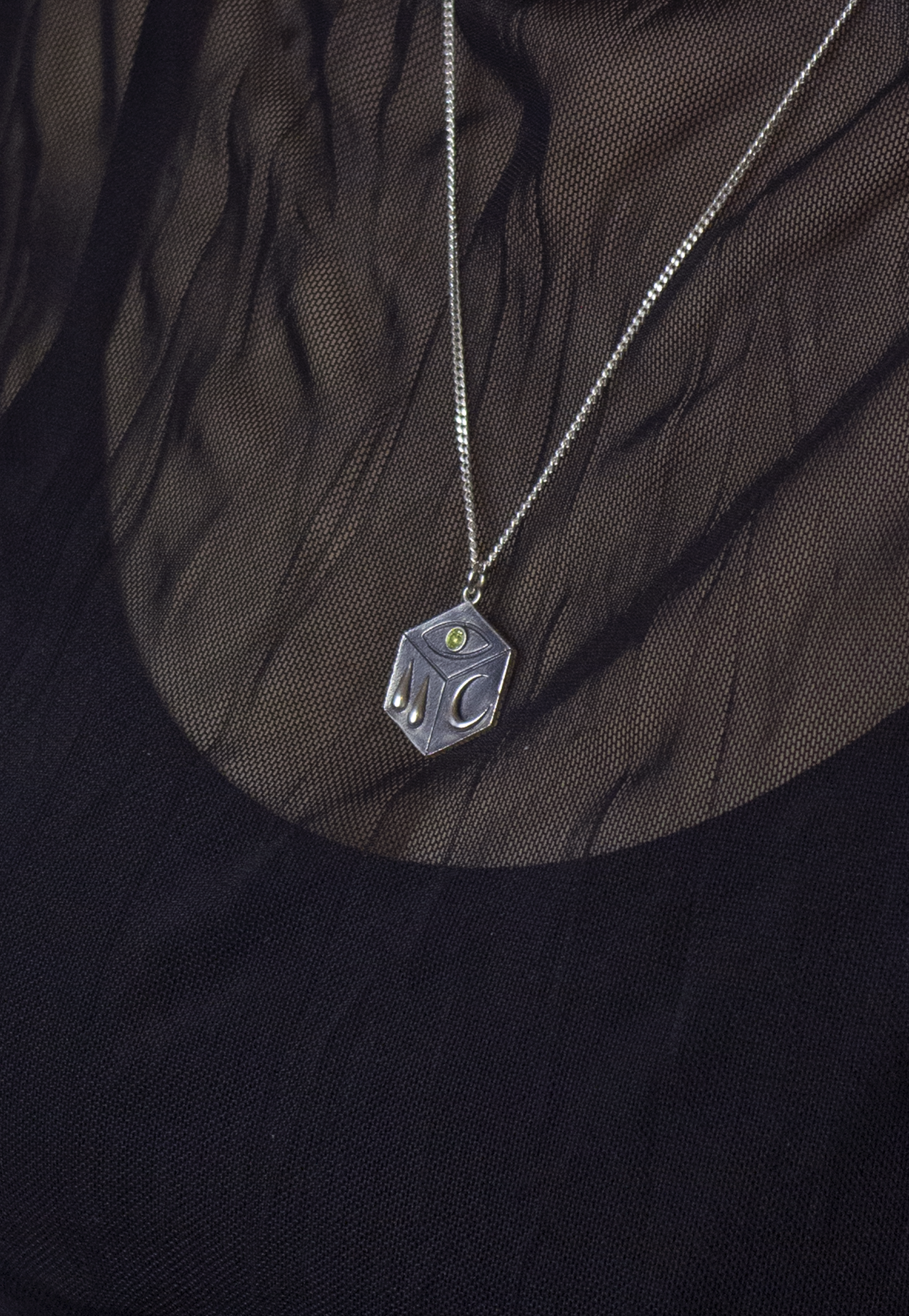 Silver necklace with a cube-shaped pendant featuring the letters 'D' and 'C' and a small green gem, worn over black fabric with sheer, patterned overlay.