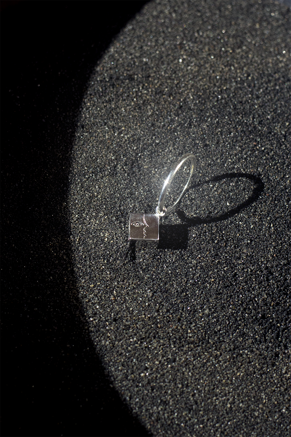 A silver ring with a square face featuring a design of a face, placed on black sand, casting a shadow.