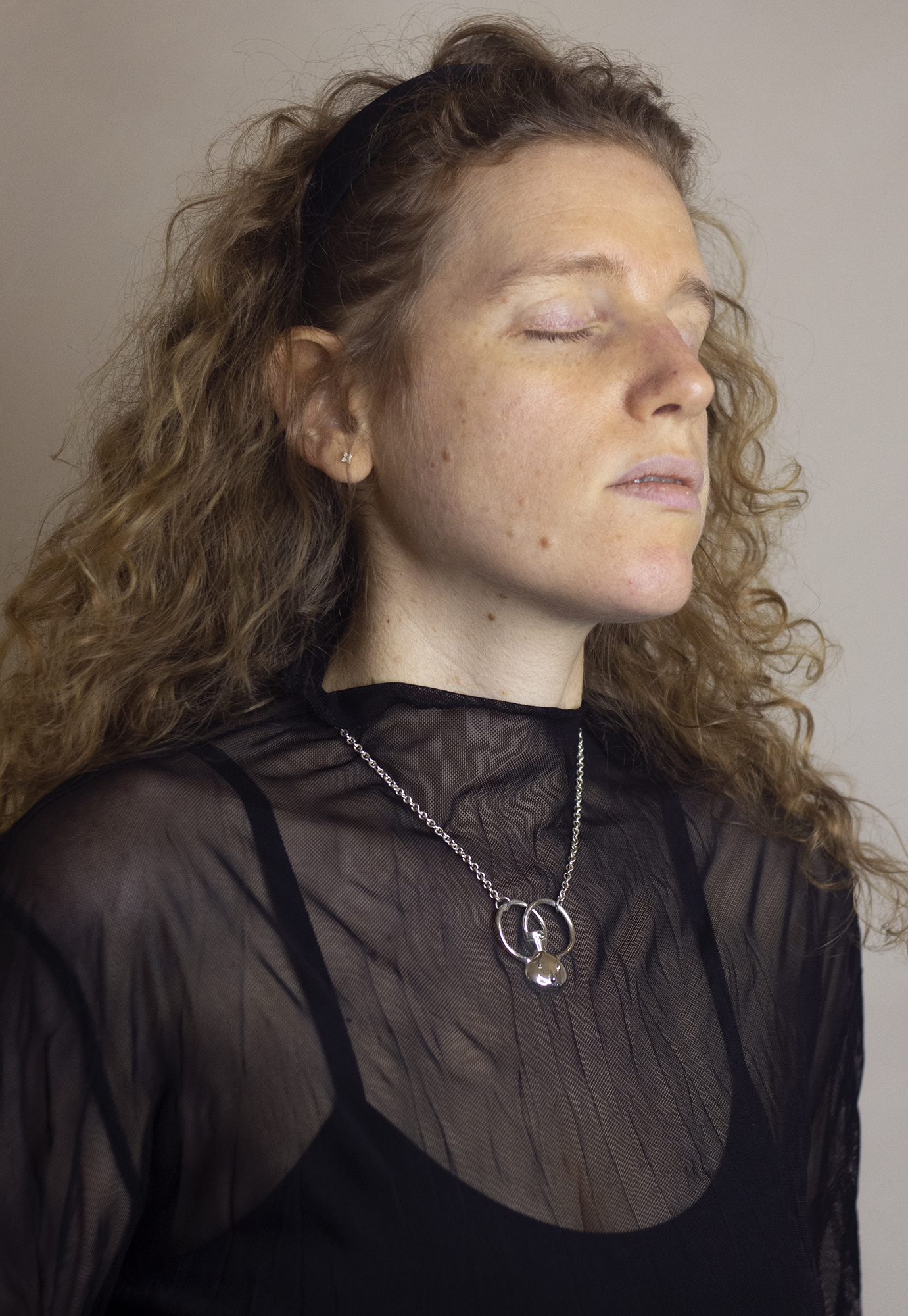 A woman with curly red hair wearing a black sheer top and silver jewelry, her eyes closed and face turned slightly upward, against a plain background.