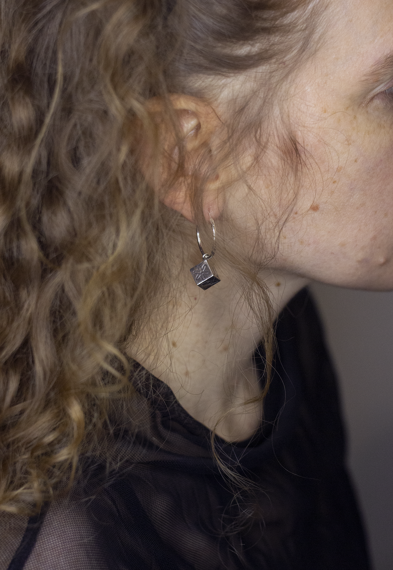 Close-up of a woman's left profile showing her curly hair, ear with silver hoop earring featuring a small cube pendant with a flower engraving, foundation makeup, and freckles on her cheek.
