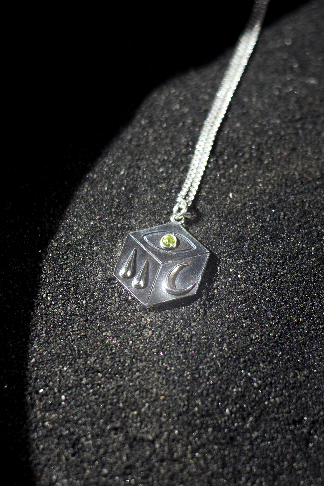A silver necklace with a pendant featuring an eye, water drops, a moon, and a green gemstone, resting on black textured surface.