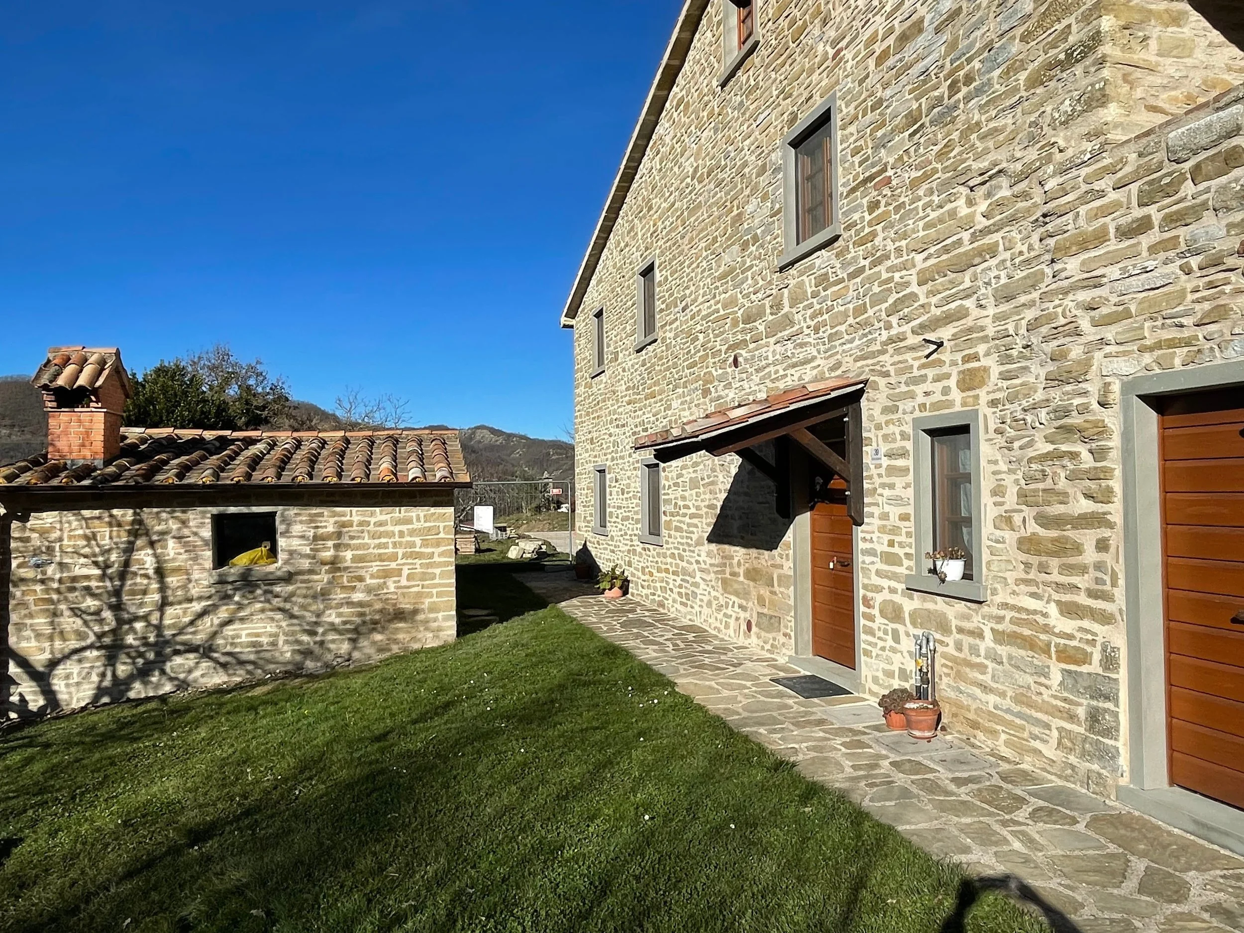 Stone house with wooden door, five windows, and a small overhang. Adjacent smaller stone structure with tiled roof. Green grass and stone pathway. Clear blue sky.