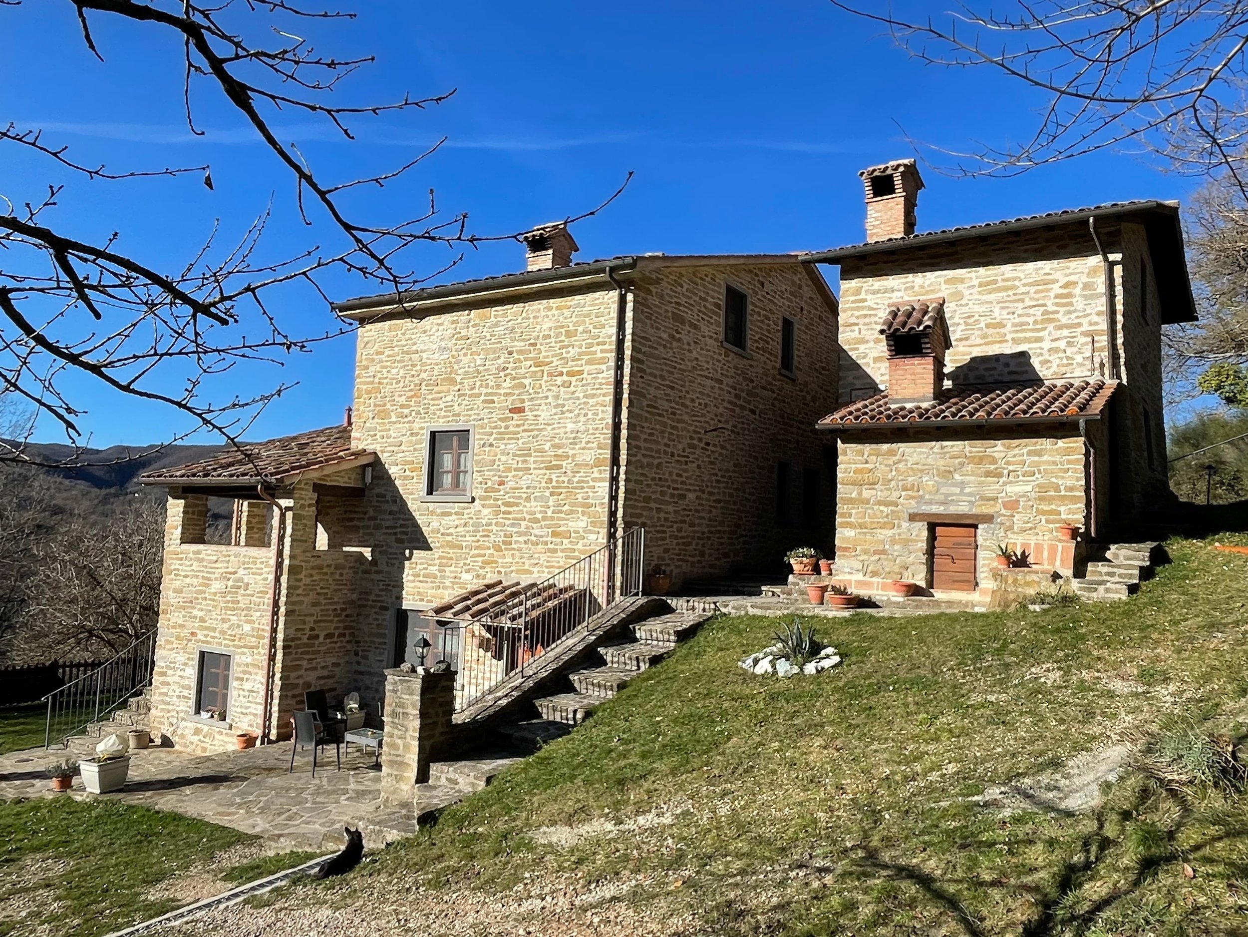 Stone house with red-tiled roof under a blue sky, surrounded by a grassy yard and leafless trees.