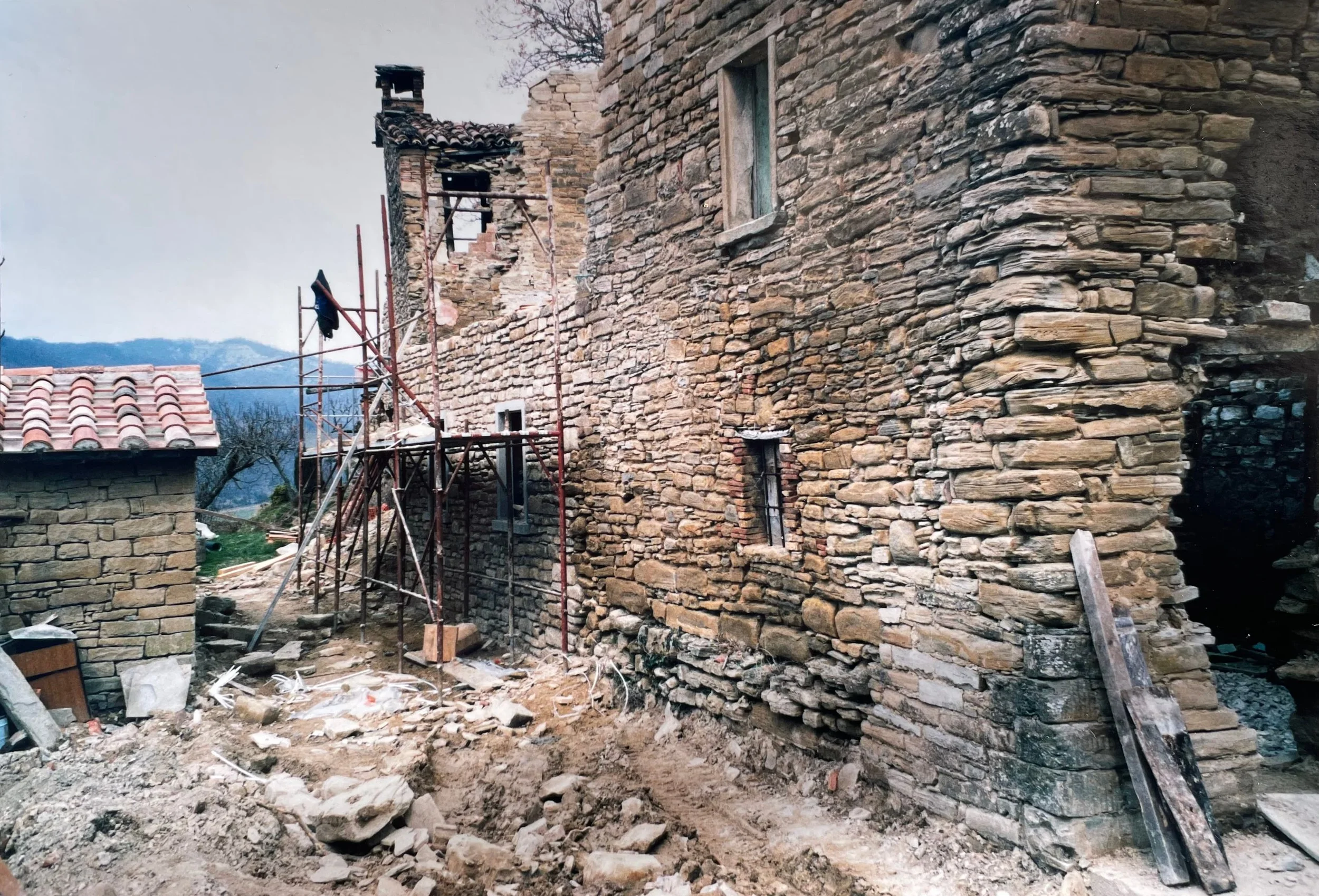 A stone building under renovation with scaffolding set up along its wall, construction debris on the ground, and small windows in the stone wall.